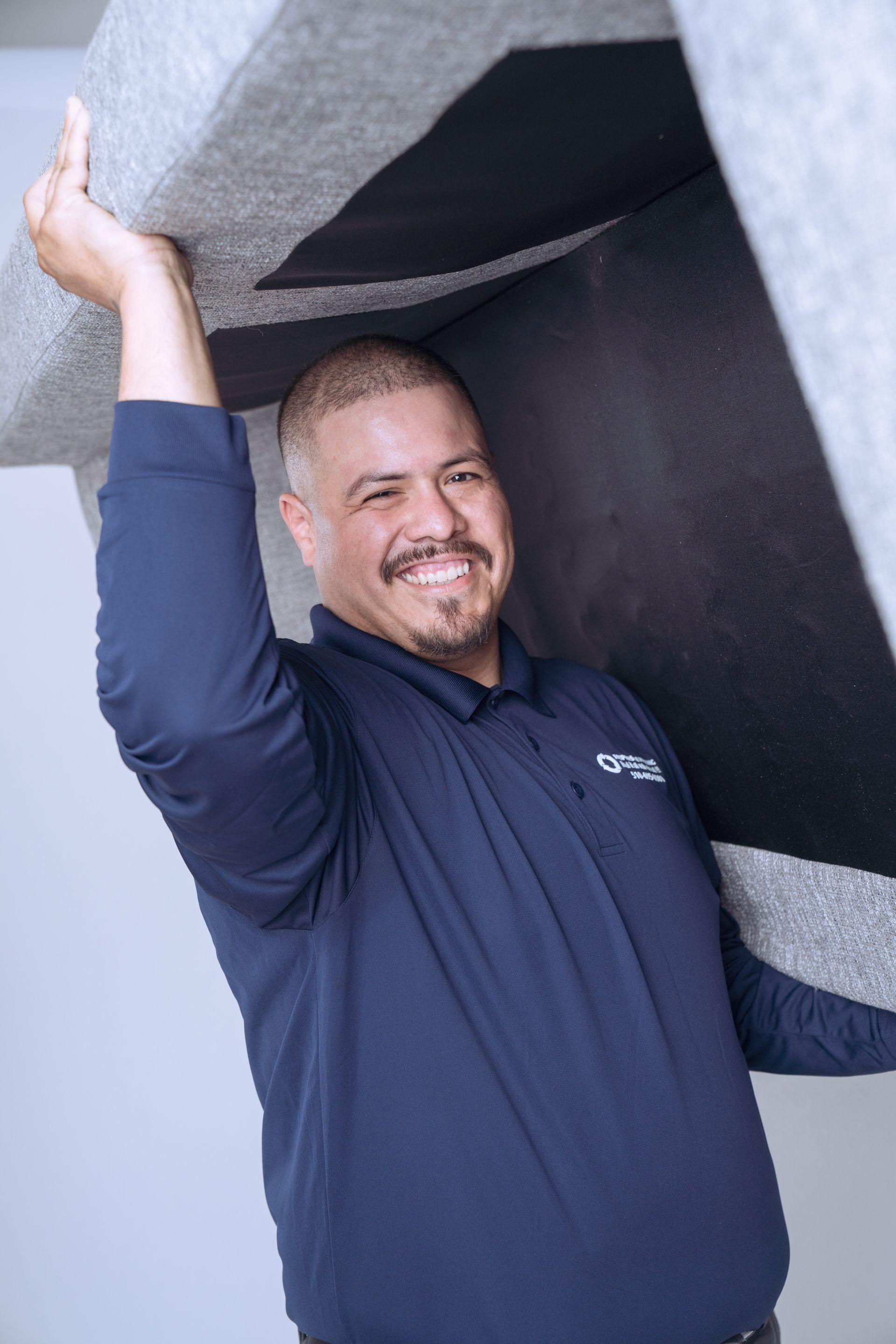 A man in a navy shirt and gray pants lifts a large beige sofa above his head.