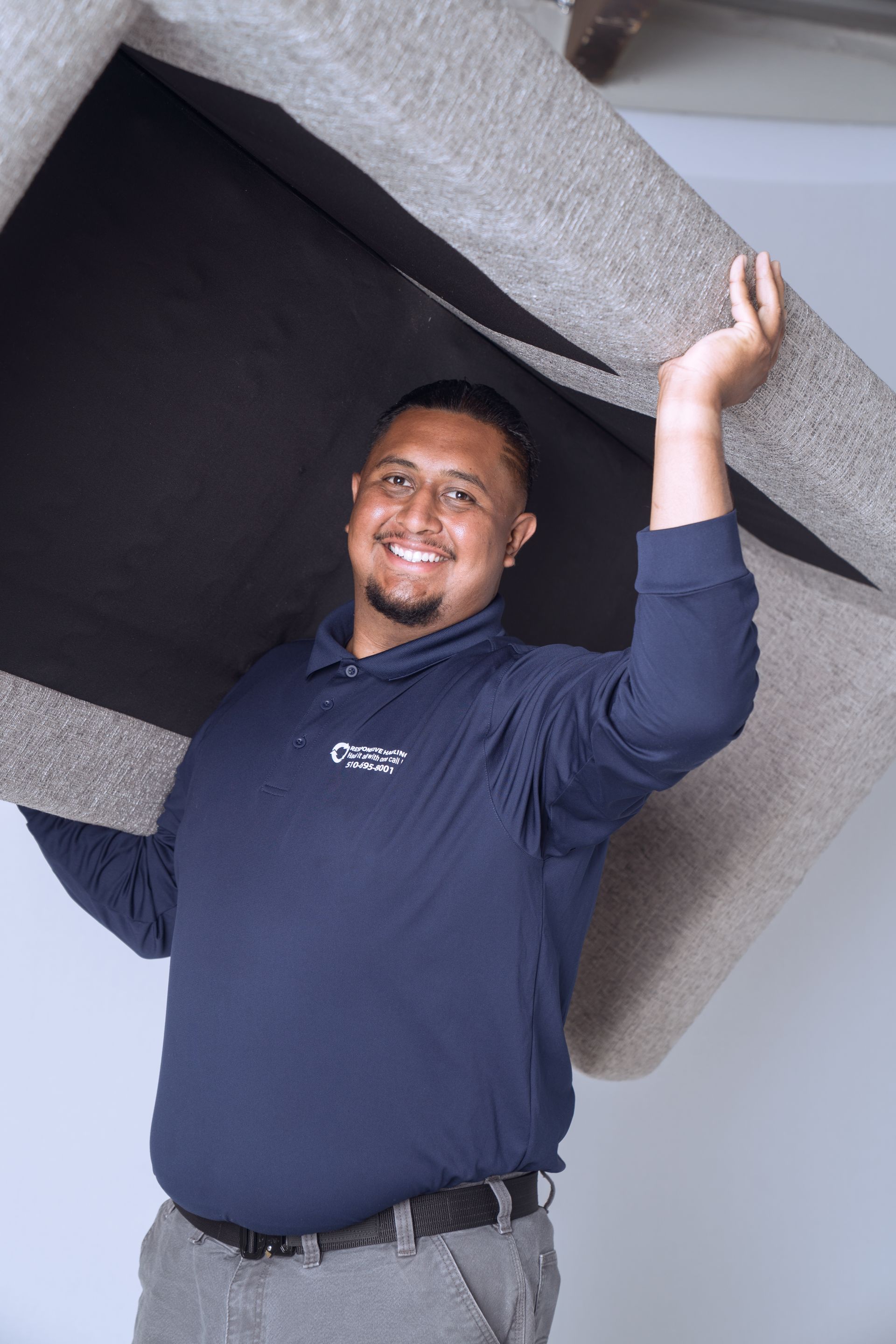A man in a navy shirt and gray pants lifts a large beige sofa above his head.