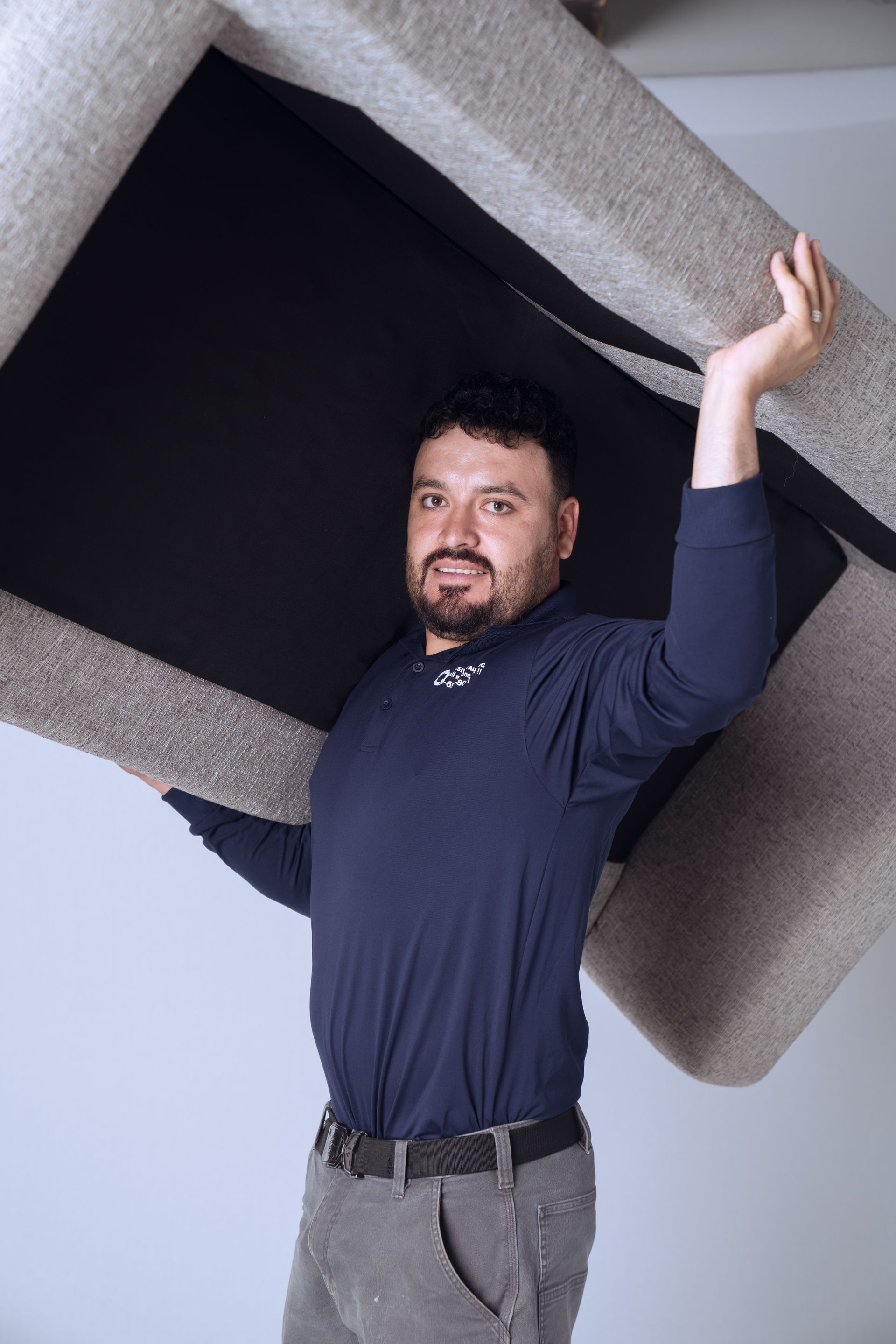 A man in a navy shirt and gray pants lifts a large beige sofa above his head.