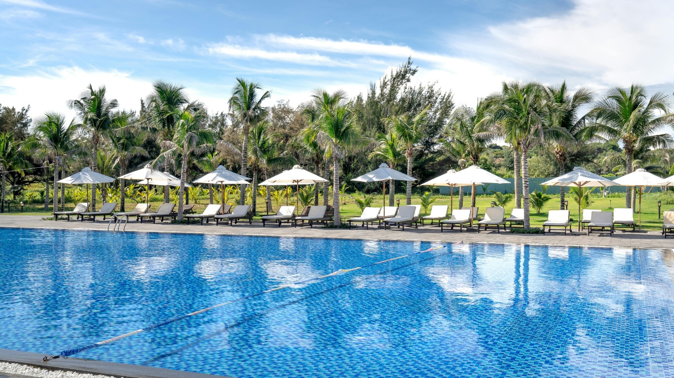 Resort pool with blue water, palm trees, and white umbrellas along the deck under a clear sky