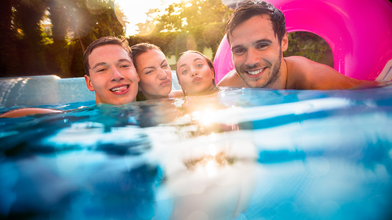 Four smiling friends in a pool, with a bright pink float in the background.