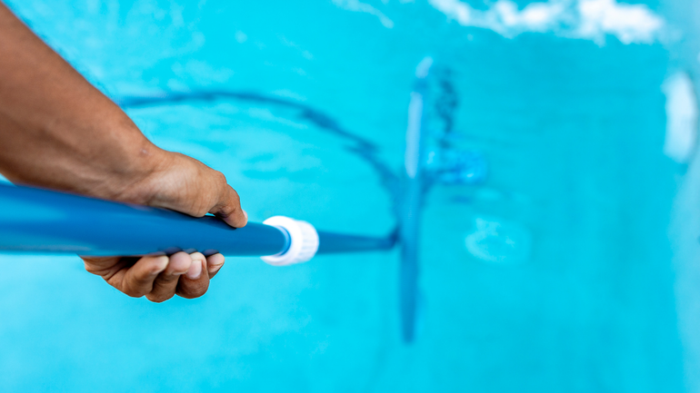 Hand holding a blue pool cleaning pole skimming the surface of bright turquoise water