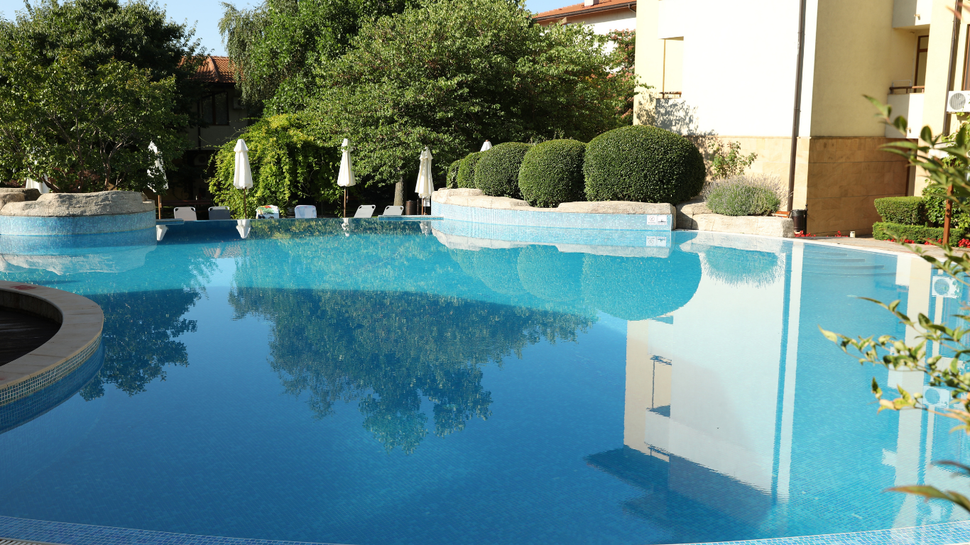 Sunny resort pool with blue water, white loungers, and manicured shrubs beside a building