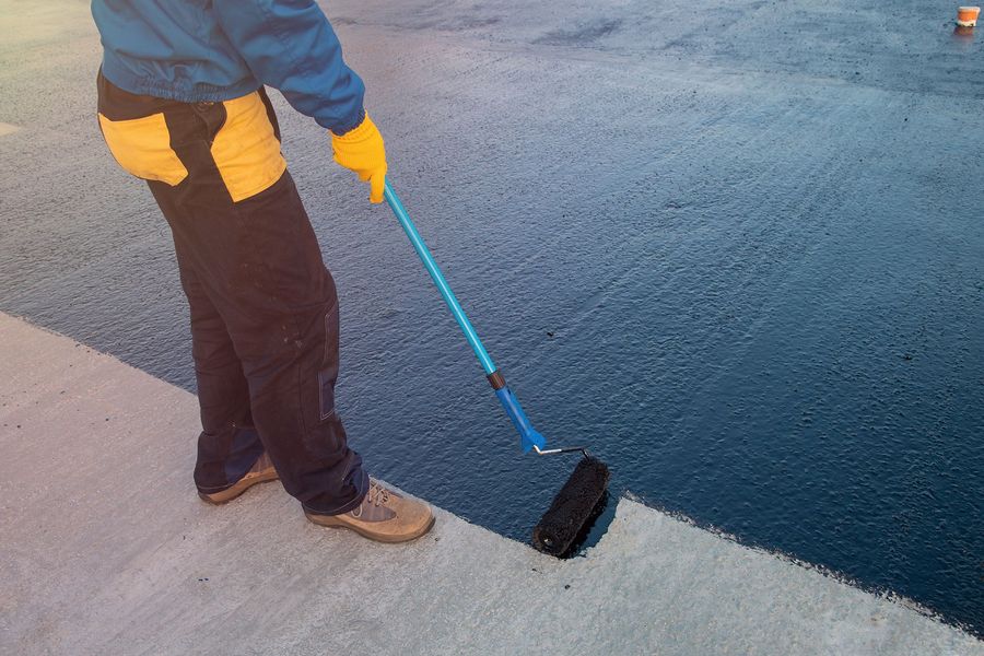 Persona aplicando sellador azul a una superficie de hormigón con un rodillo. Lleva ropa de trabajo y guantes en un entorno exterior.