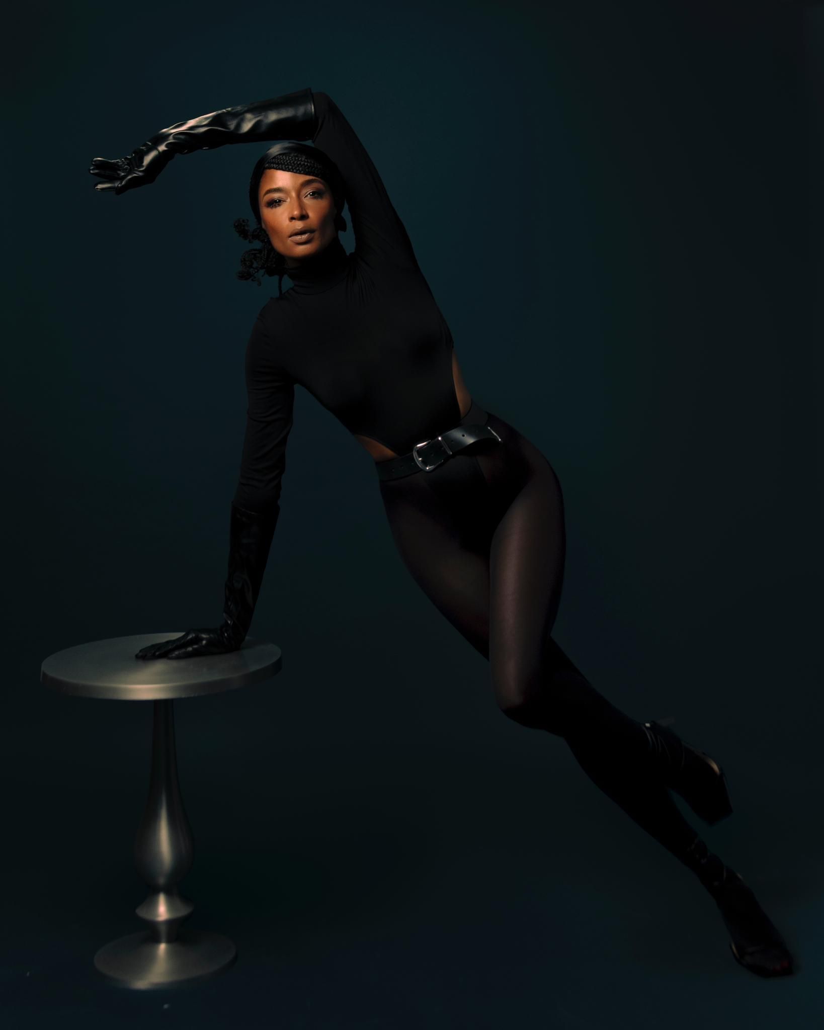 A woman in a black bodysuit and gloves is sitting on a table.