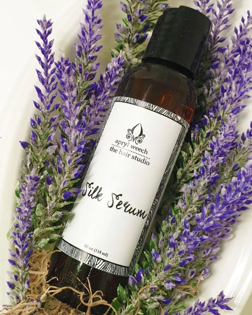 A bottle of silk serum is sitting on top of a white plate surrounded by purple flowers.