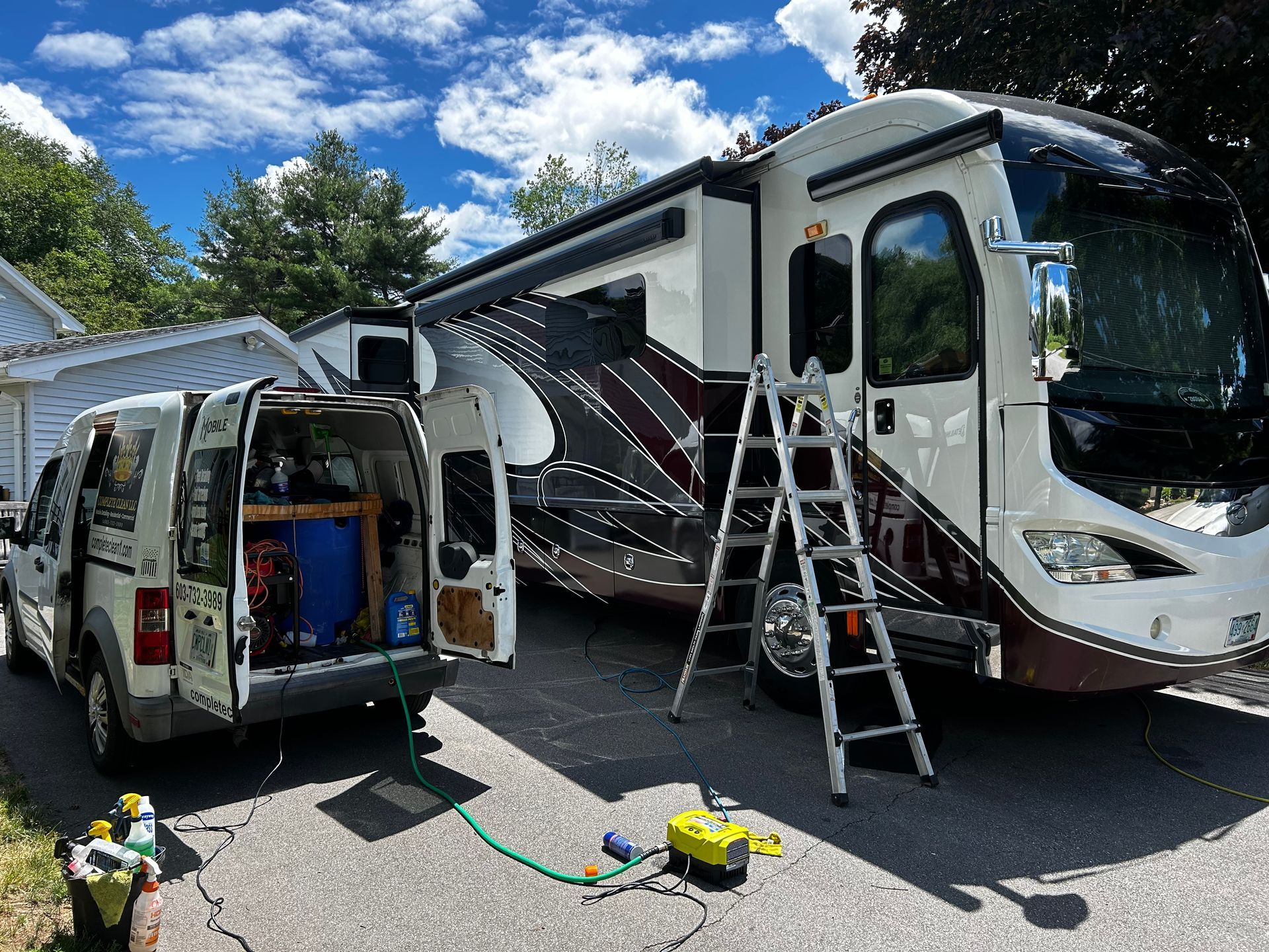 A motorhome parked in a driveway with a van connected to it for cleaning. A ladder stands near the RV.