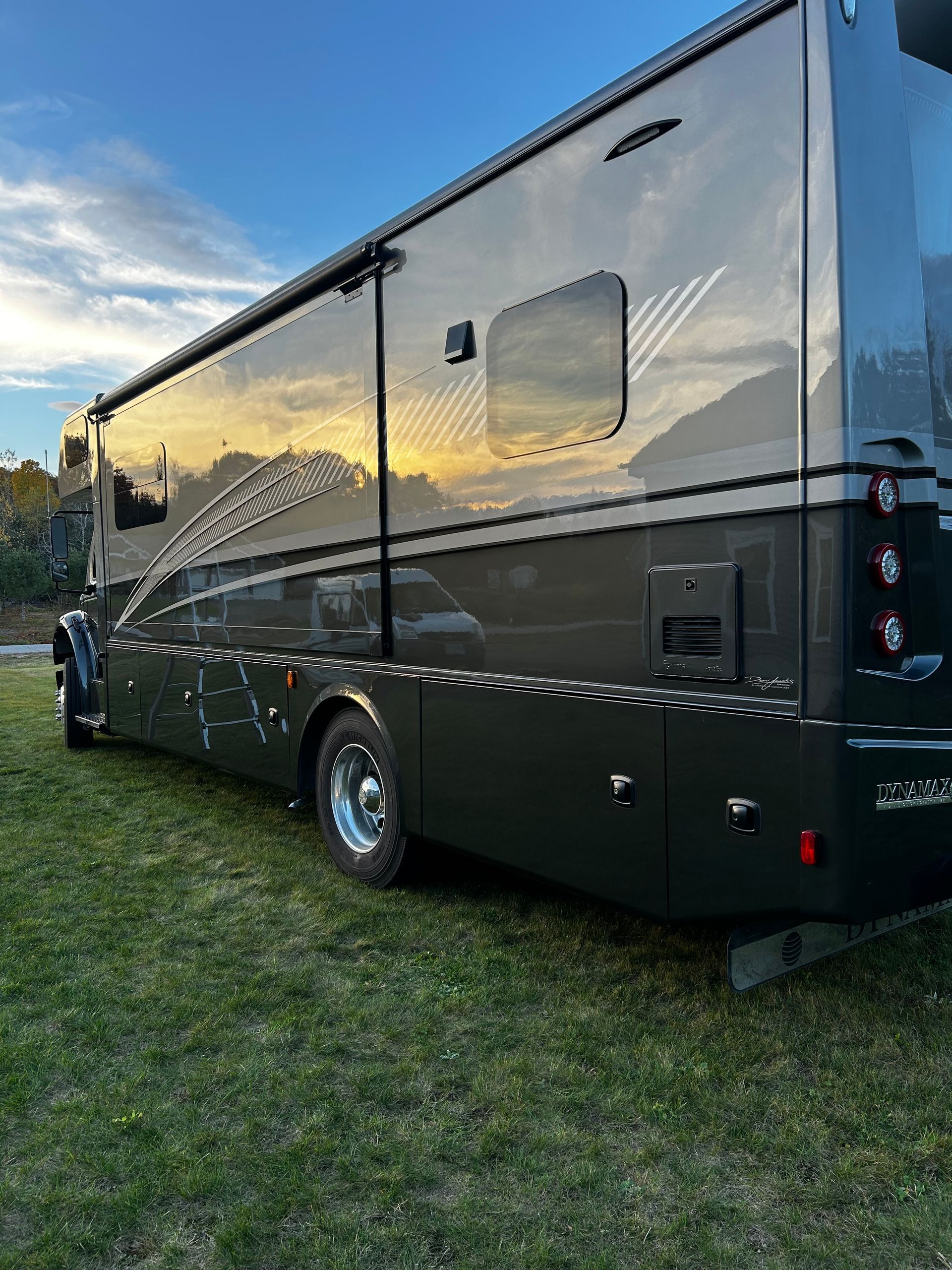 Dark gray RV parked on grass with the sky reflecting in its windows. The awning is extended.