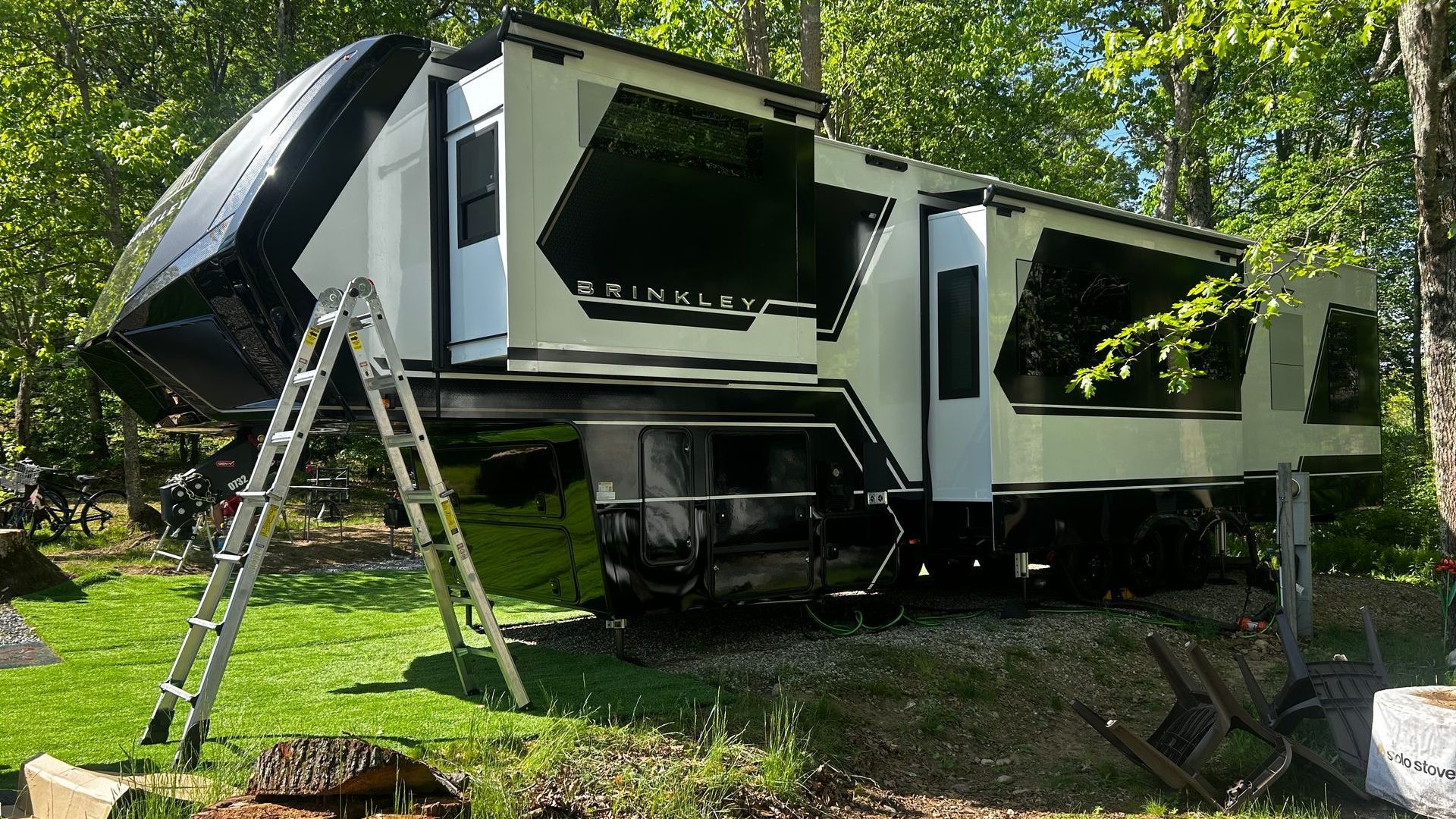 Modern black and white RV parked on grass with a ladder leaning against it, surrounded by trees.