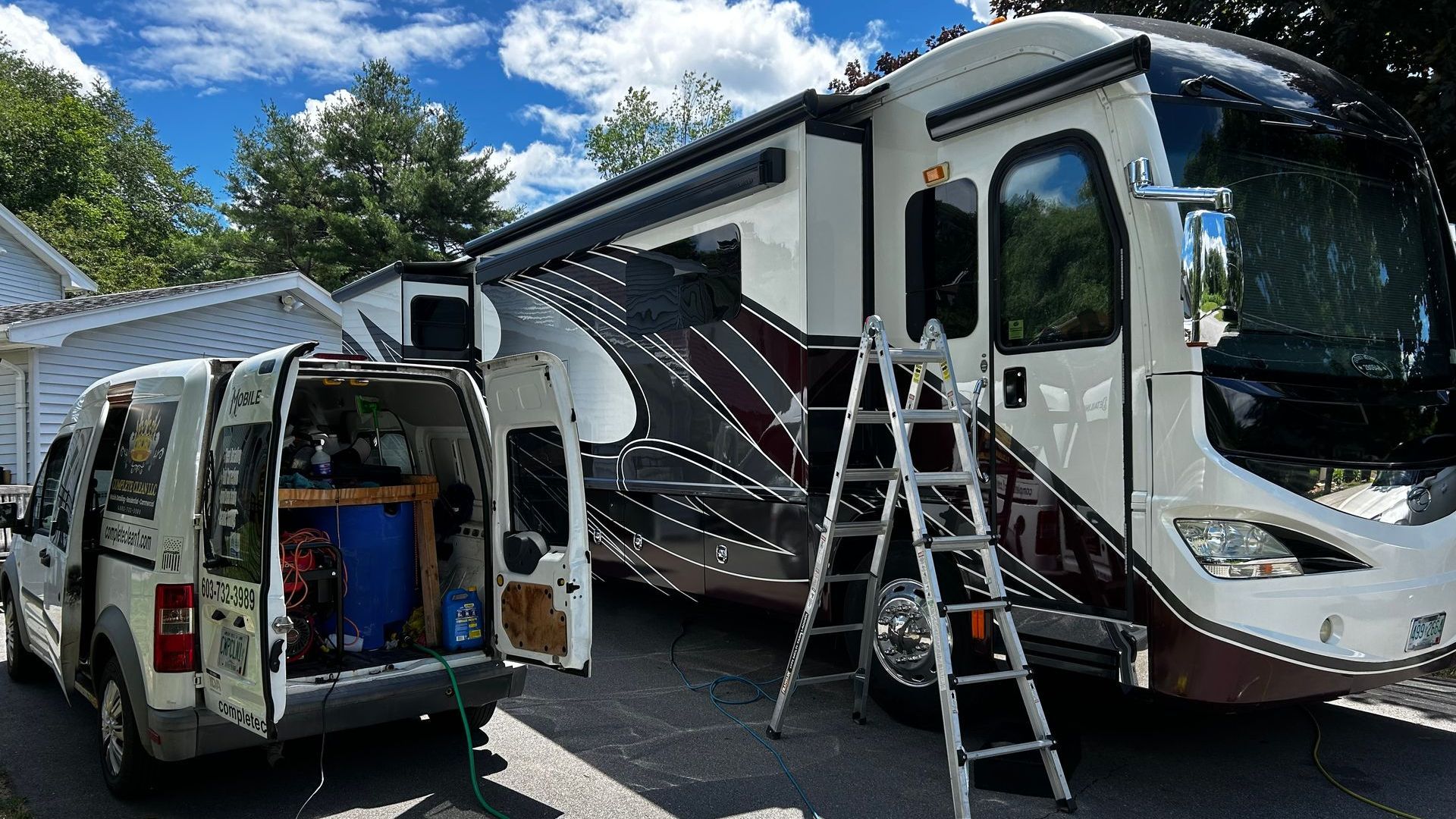 A white and maroon RV parked next to a white van with its doors open. A ladder is leaning against the RV.