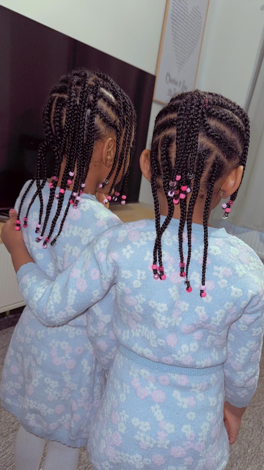 Two young girls, wearing matching blue floral sweaters, embrace. Braided hairstyles with pink beads.