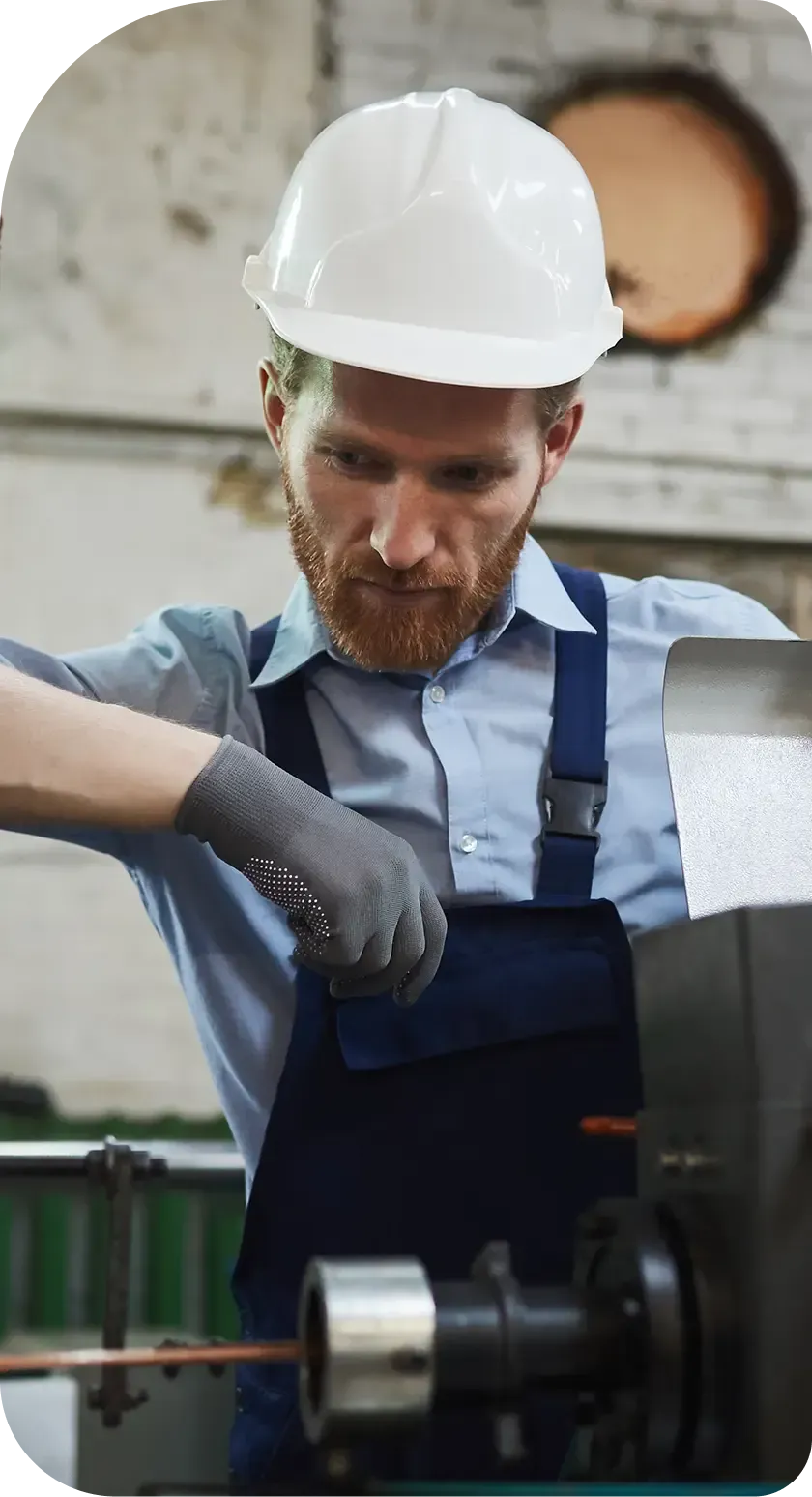 A worker in a white hard hat and blue coveralls operates industrial machinery in a workshop, focusing on the task.