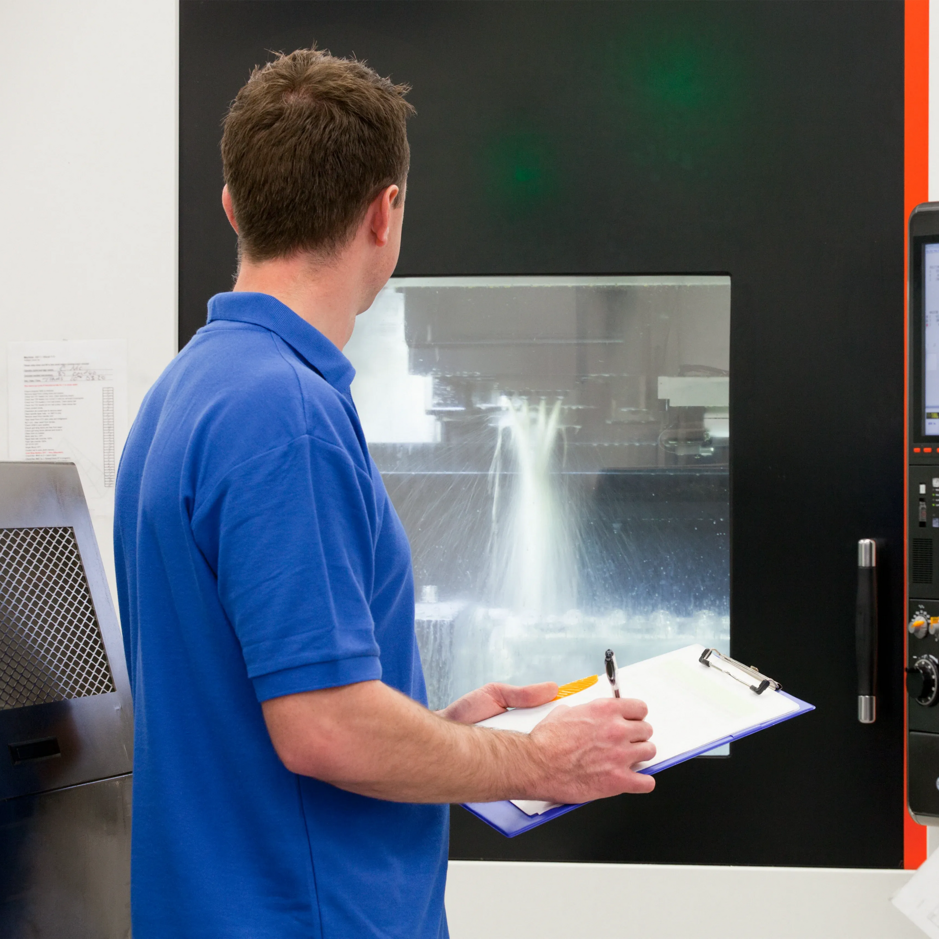 A person in a blue shirt holds a clipboard and pen while monitoring a machine operating with liquid coolant.