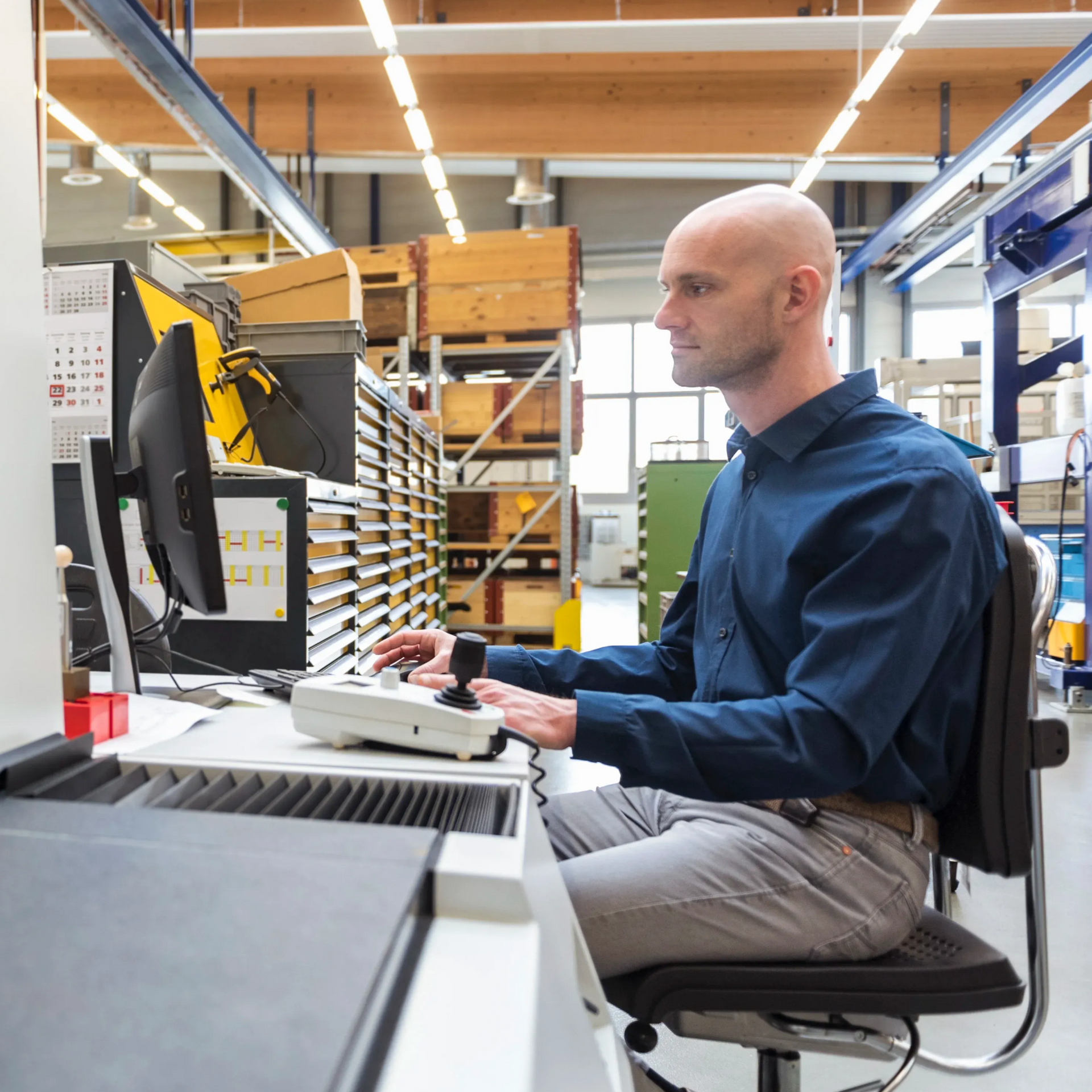 A professional operates a control console with a joystick and computer screen in an industrial factory setting.