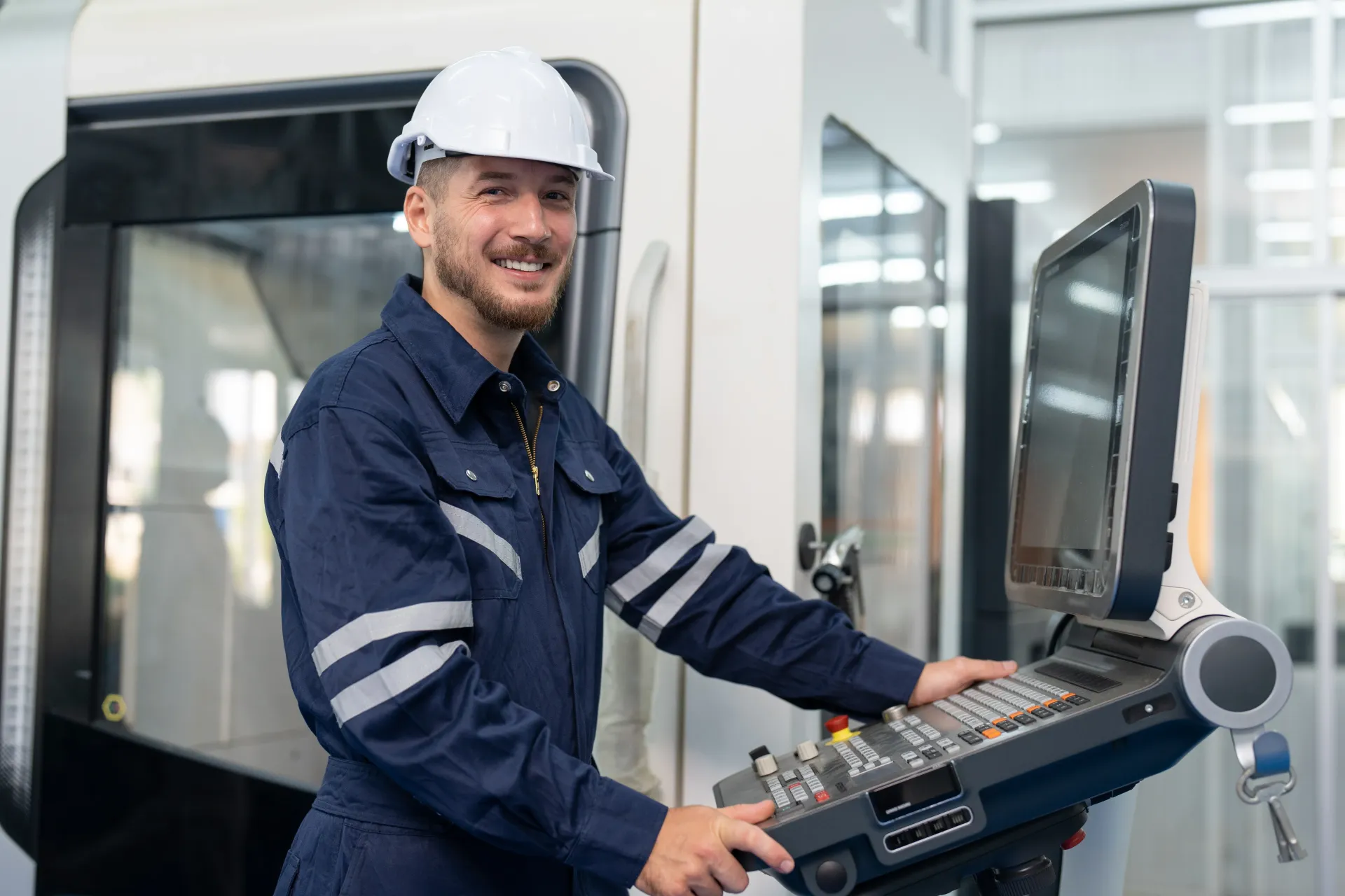 A smiling factory worker in a hard hat and blue uniform operating the control panel of a large industrial machine.