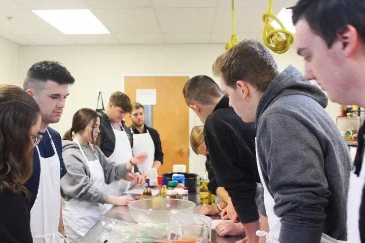 A group of people wearing white aprons gathers around a stainless steel counter in a kitchen, observing food preparation.