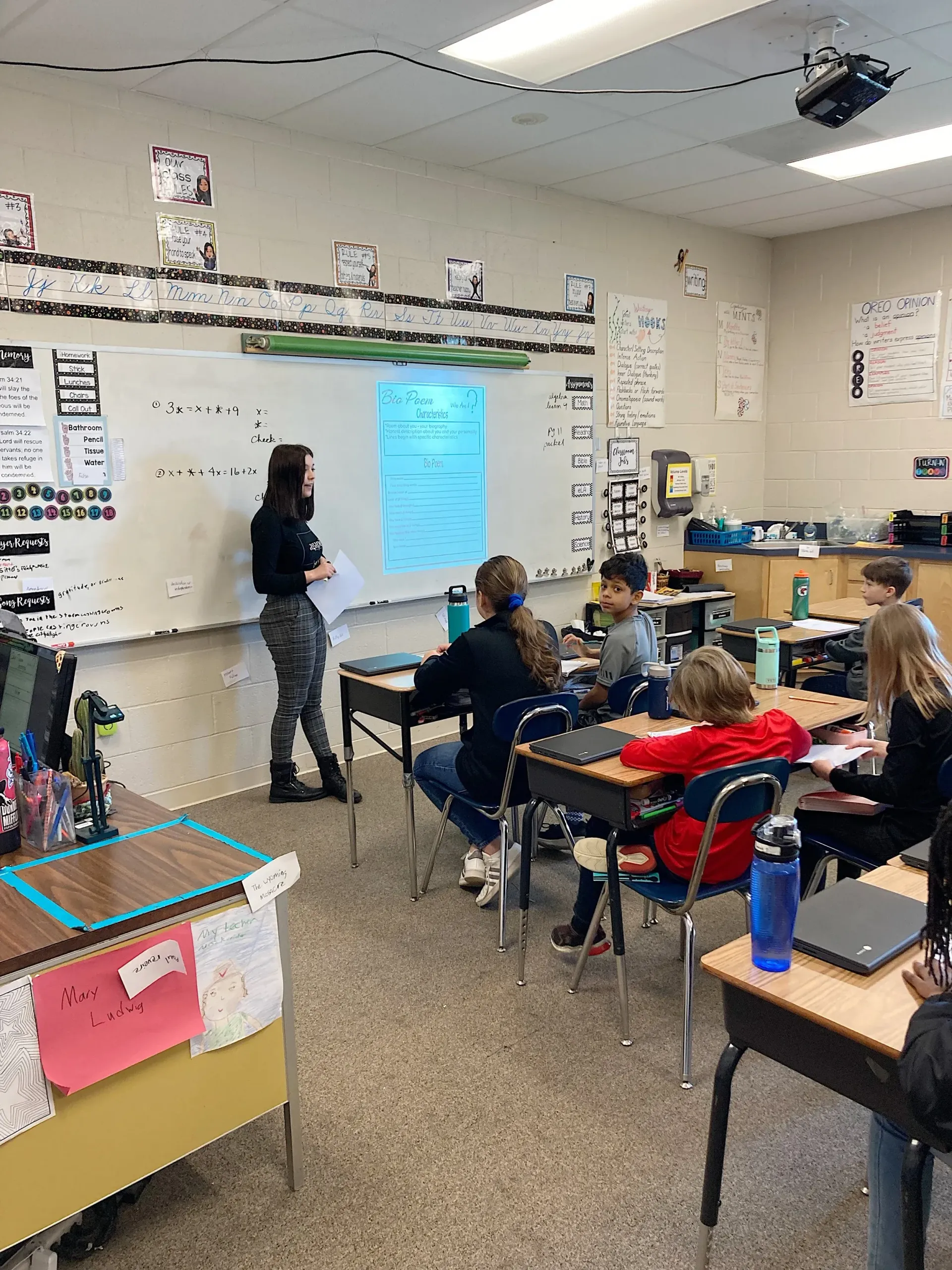 Teacher at whiteboard; students in desks. Classroom setting, blue paper on the board, laptop on desk.