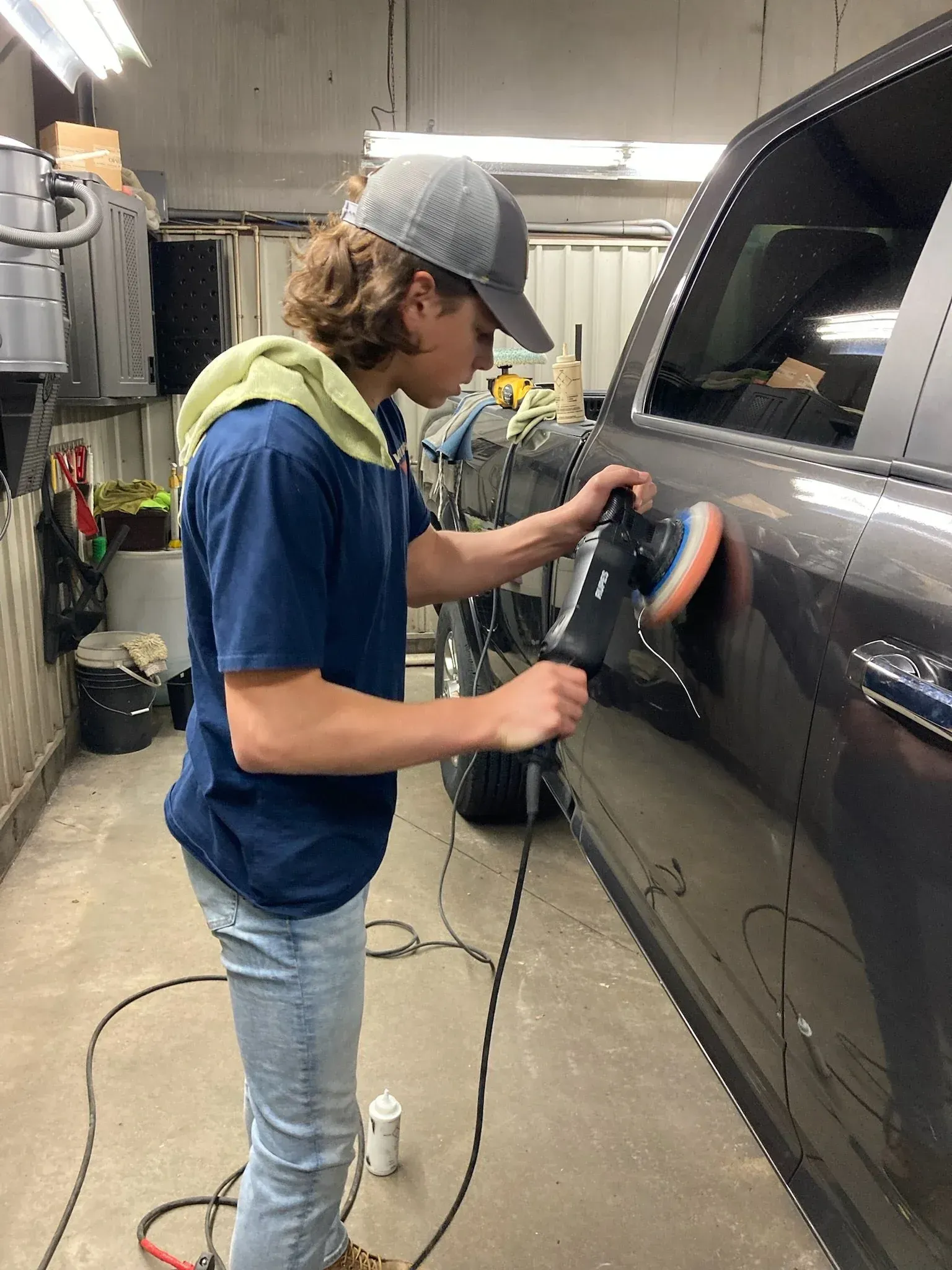 Person polishing a dark car's side panel with a machine in a garage setting.