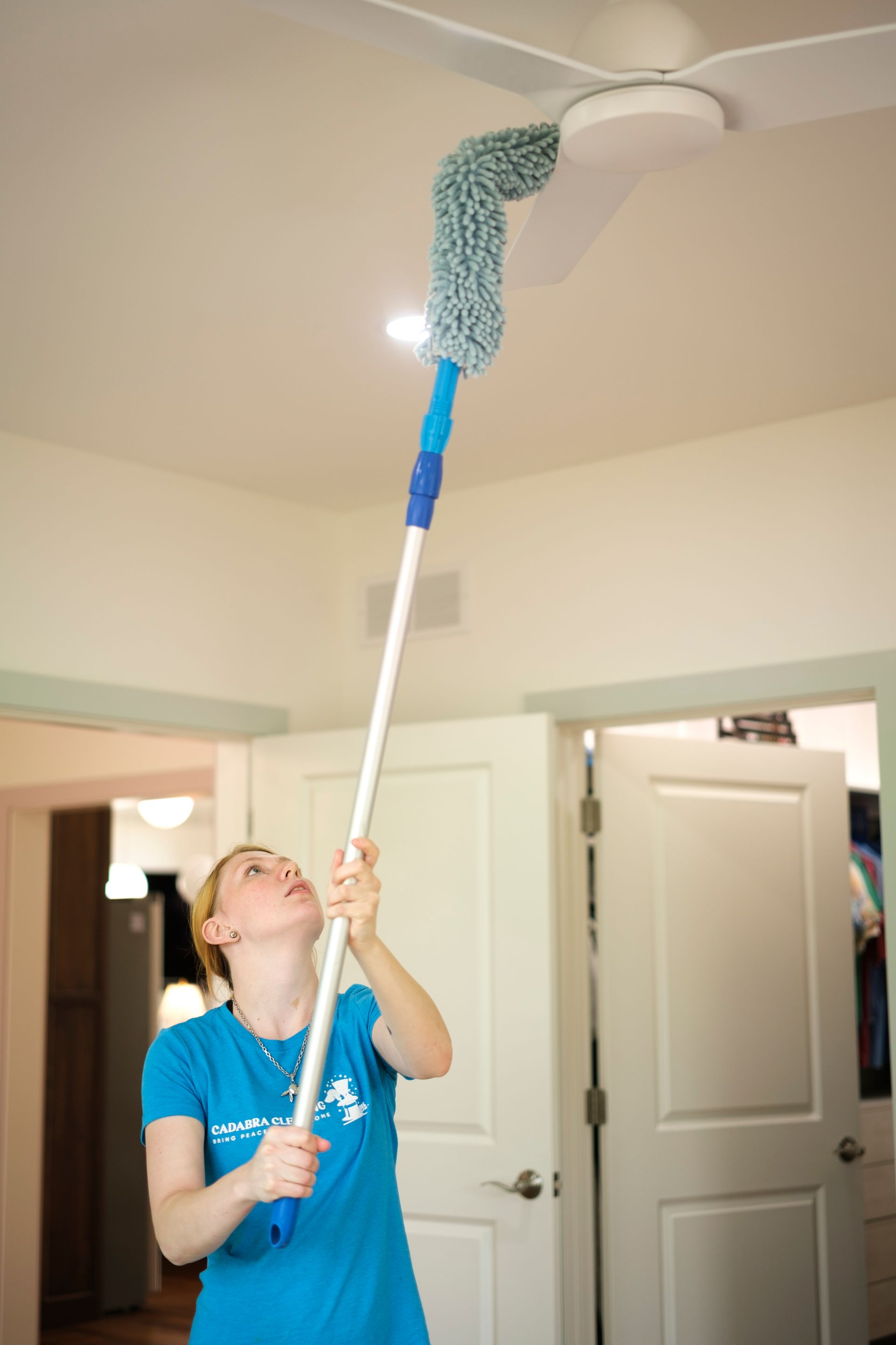 Woman dusting a ceiling fan with a long-handled duster in a room with white doors.