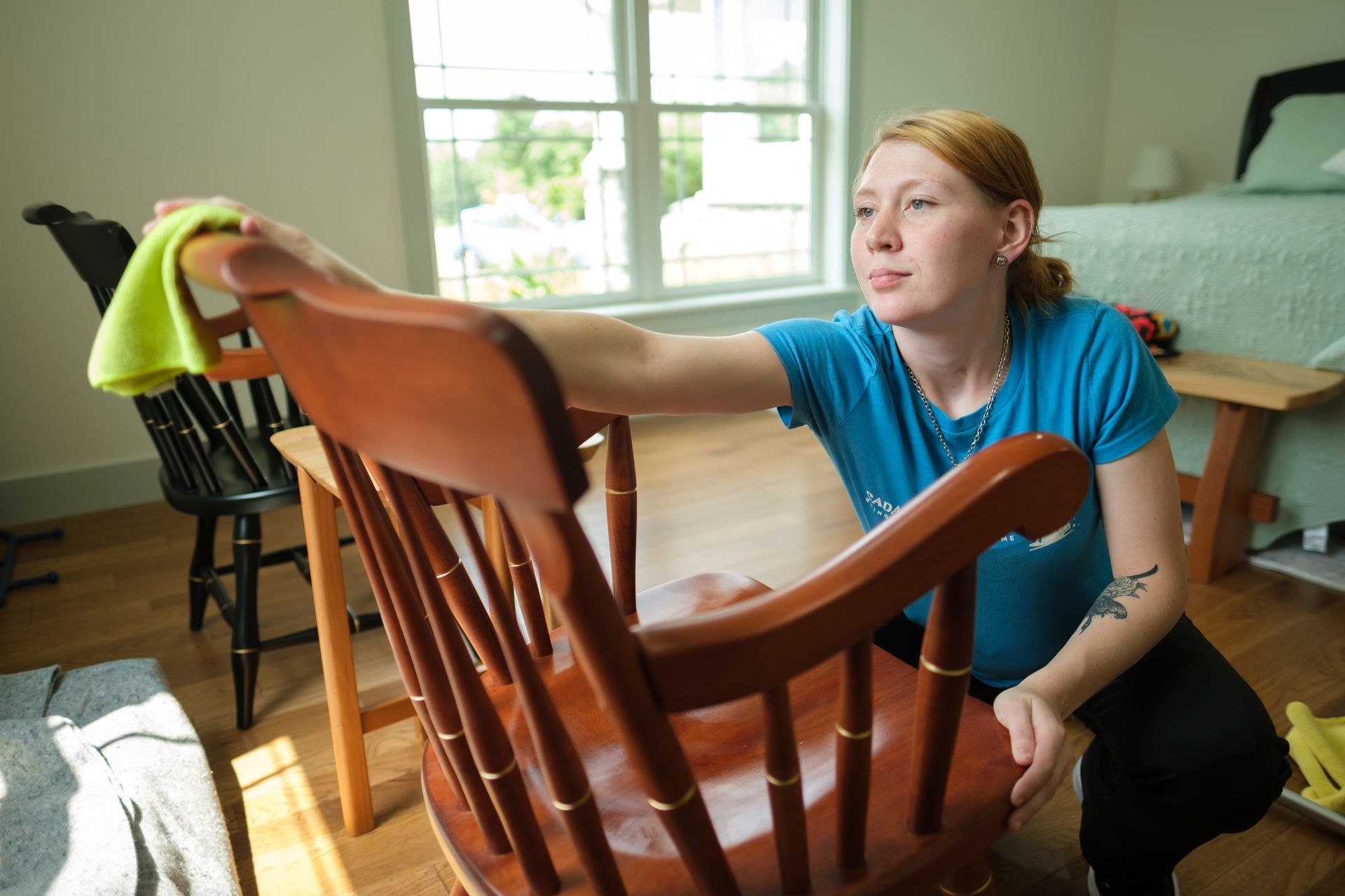 Woman cleans a wooden chair with a yellow cloth in a sunlit room.