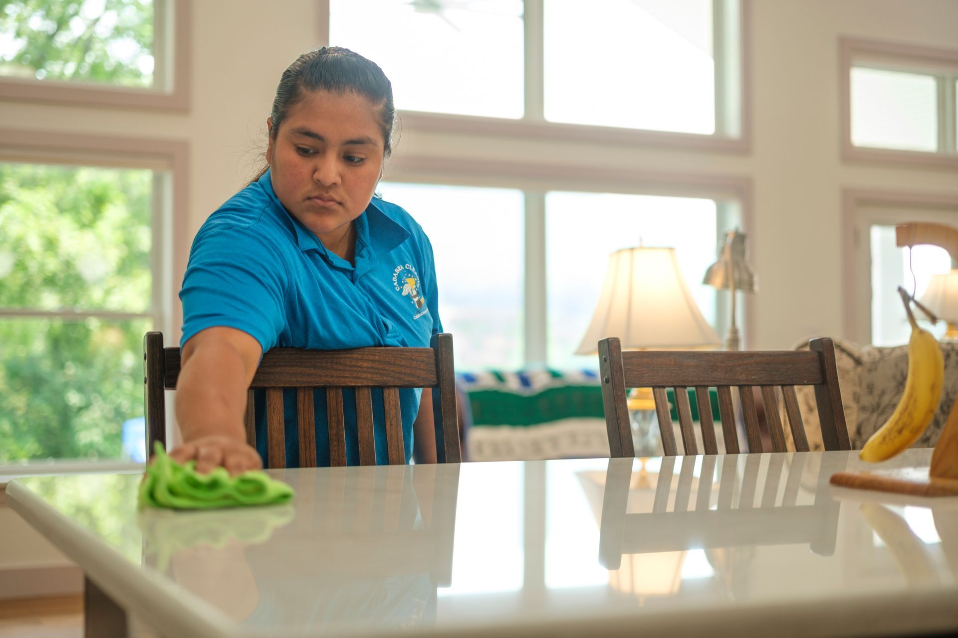 Woman in blue shirt wiping a white countertop in a bright kitchen.