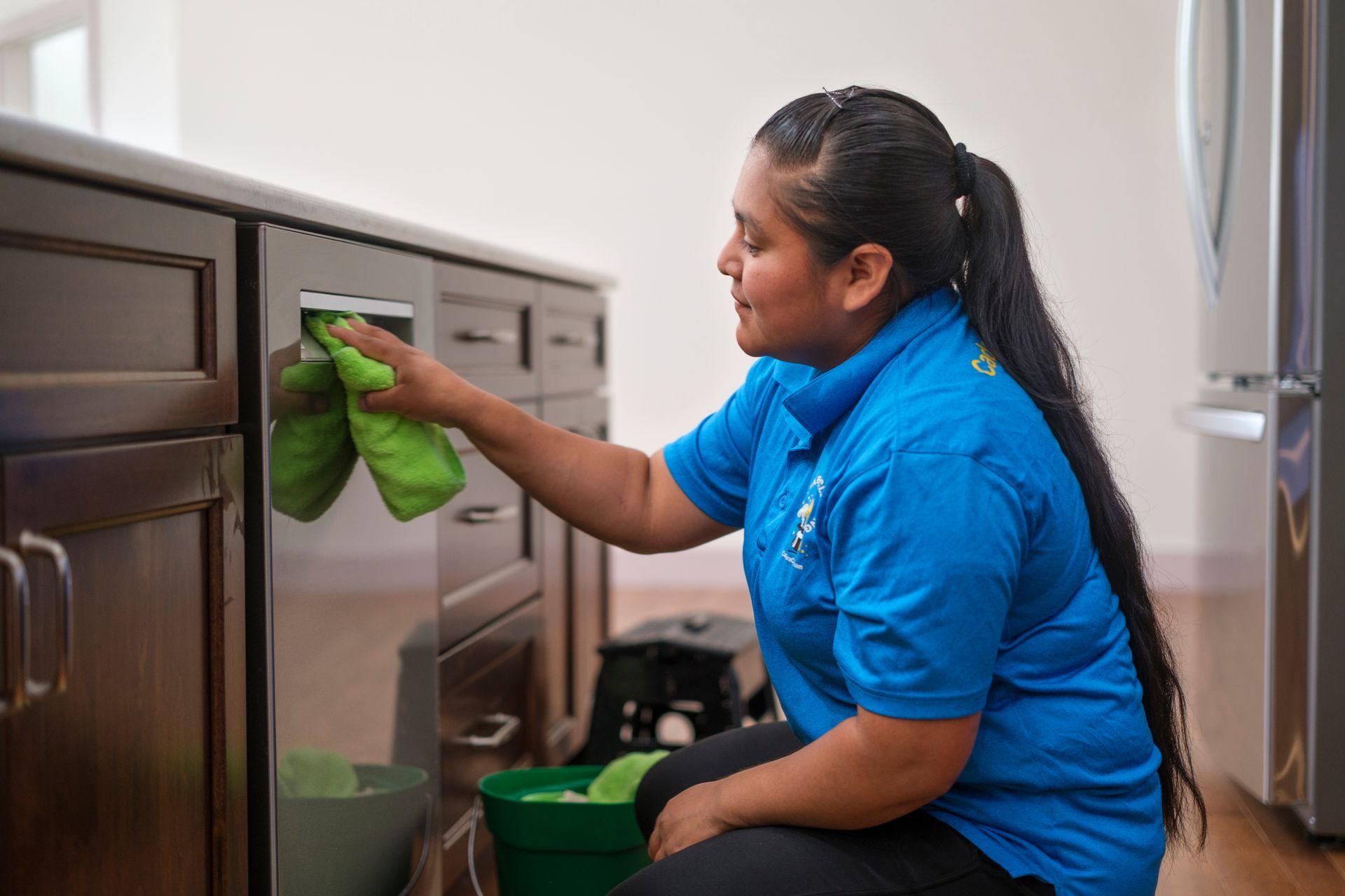 Woman in blue shirt cleans kitchen cabinet with green cloth.