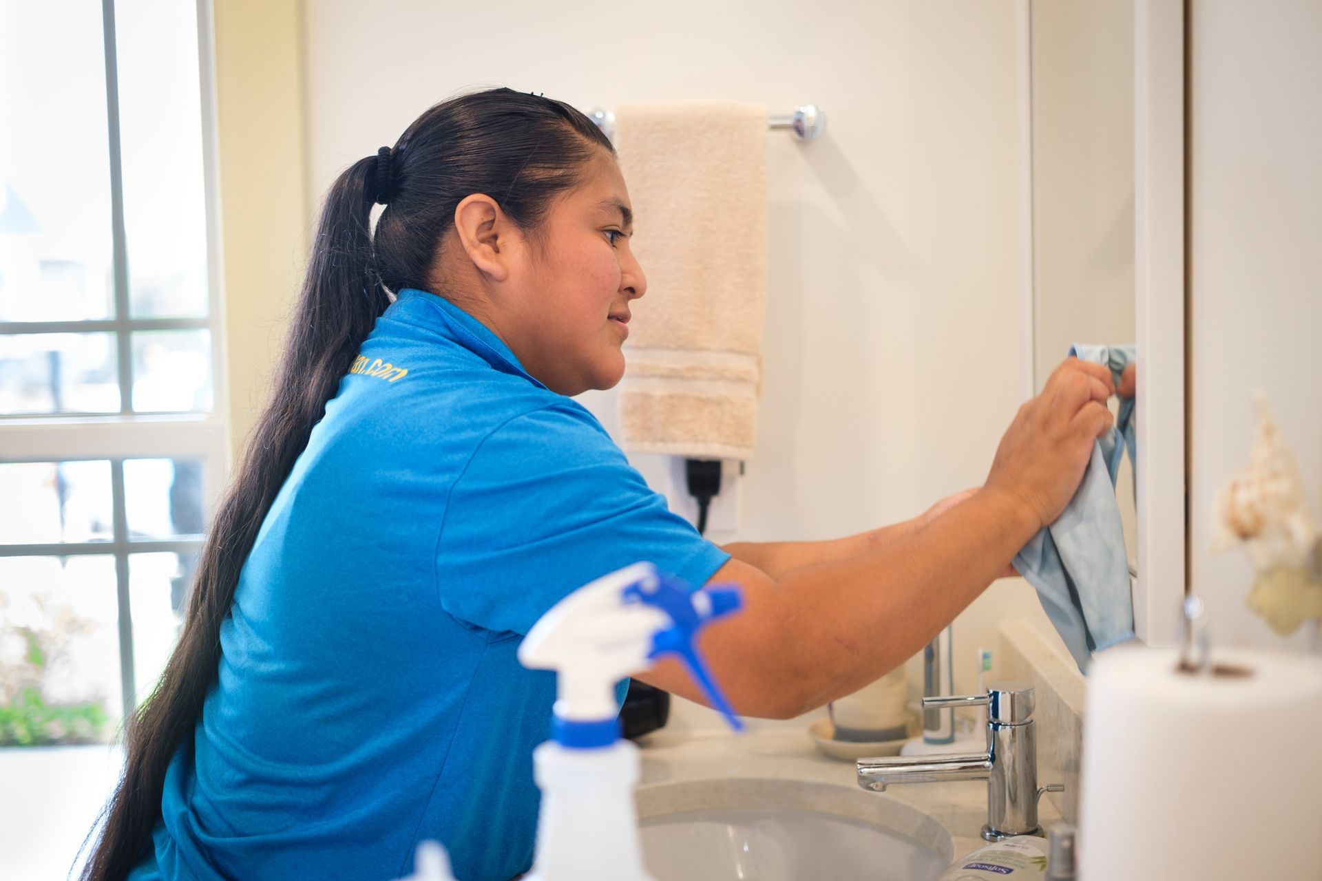 Person cleaning a bathroom mirror. They wear a blue shirt, have long black hair, and are using a cloth and spray bottle.