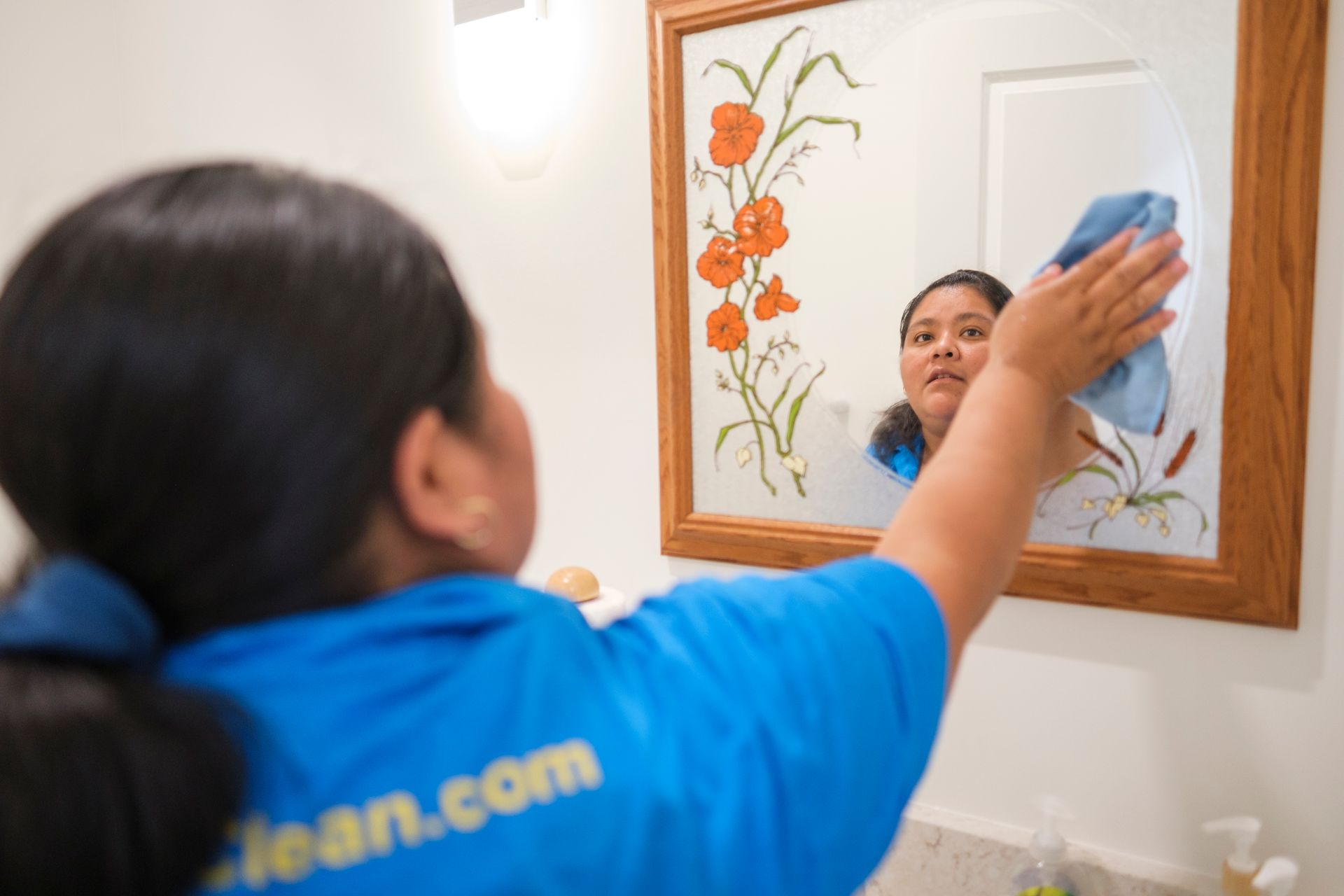 Woman cleaning a mirror in a bathroom; wearing blue shirt, reflection visible.