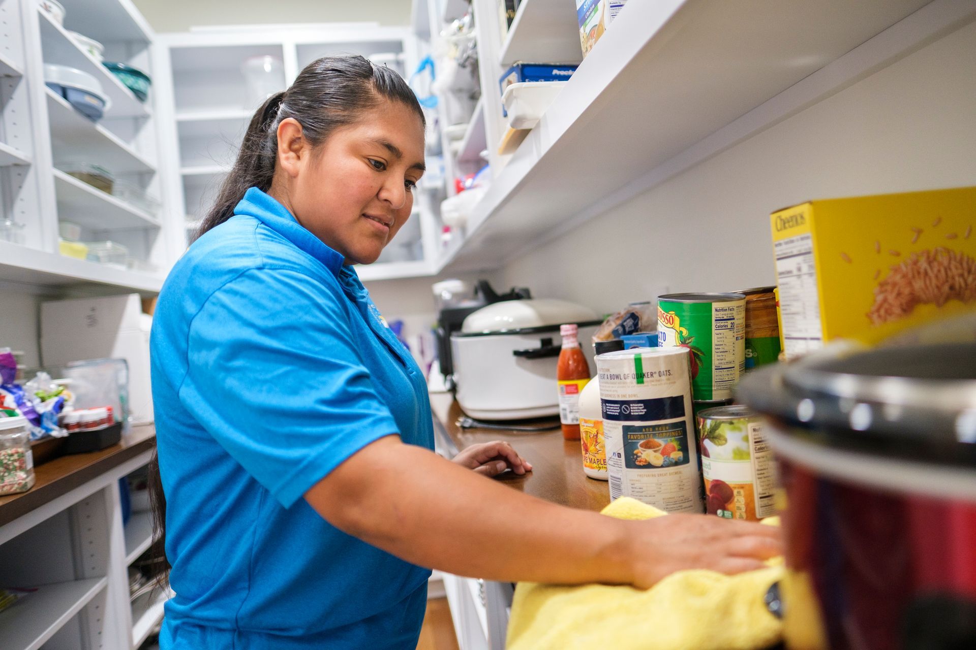 Woman in blue shirt wiping a pantry shelf with a yellow cloth.