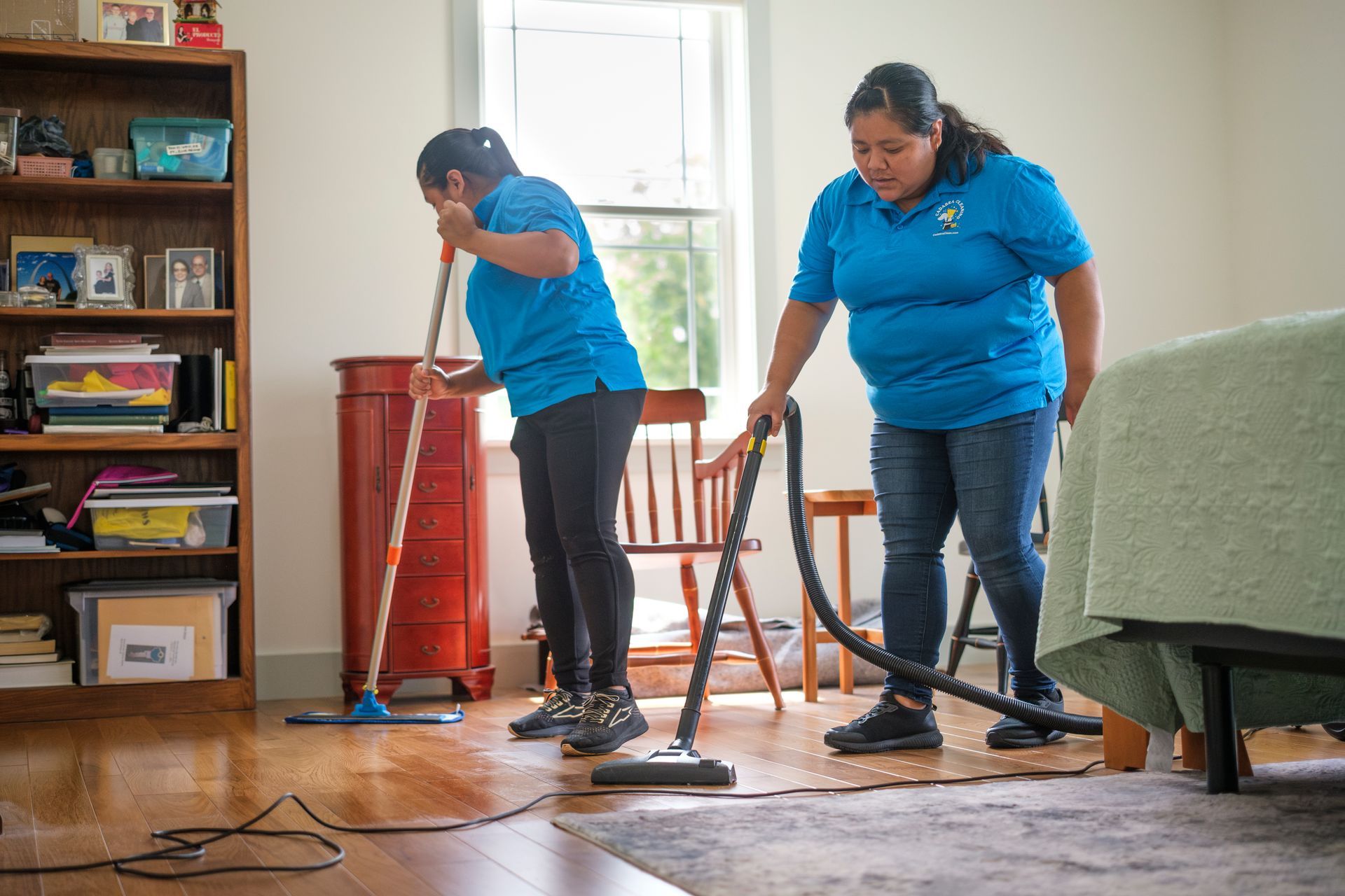 Two people cleaning a room: one mopping, the other vacuuming. Hardwood floor, bookshelf, bed, window.