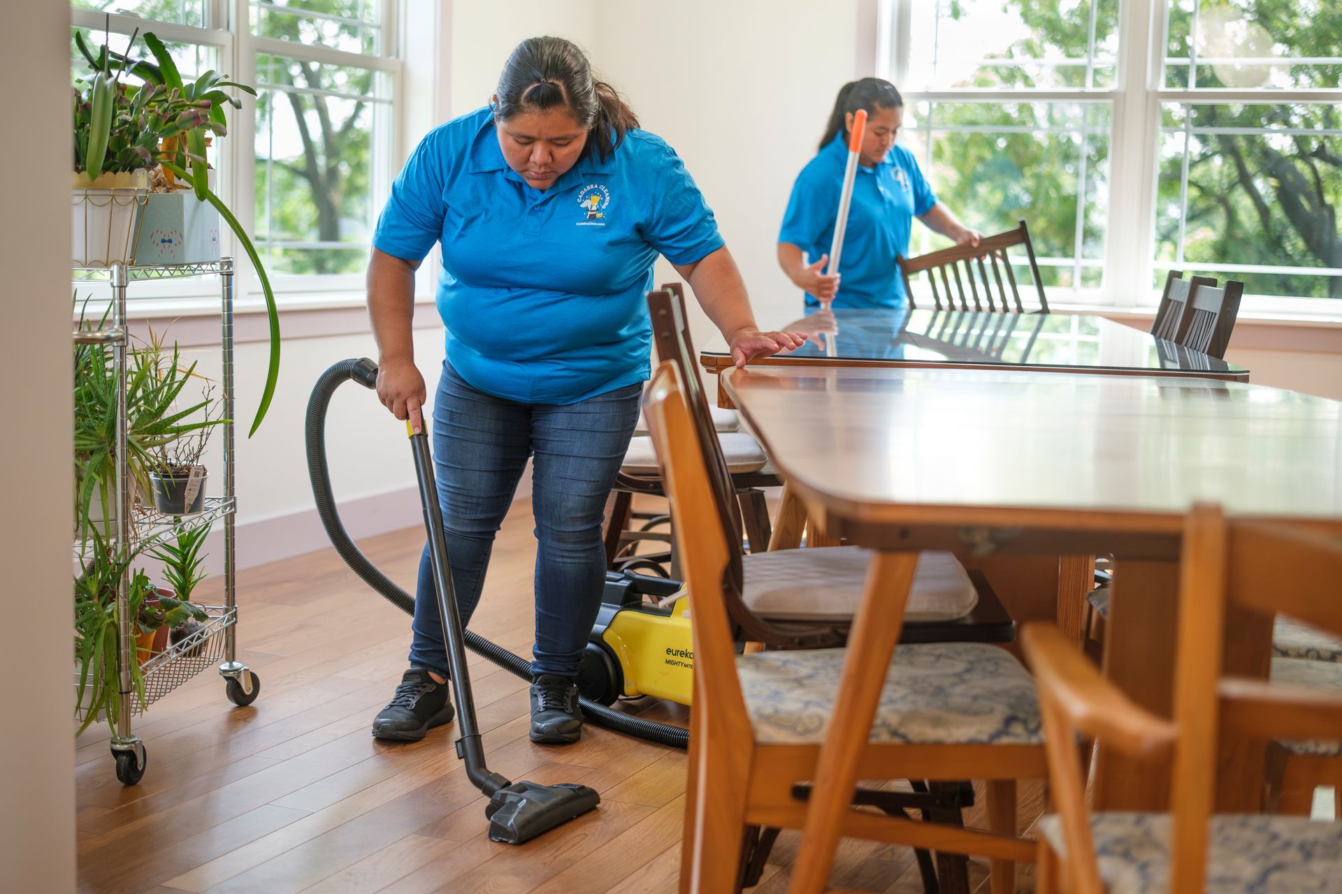 Two people cleaning a dining room. One vacuums the floor, the other dusts the table. Bright, sunny room.