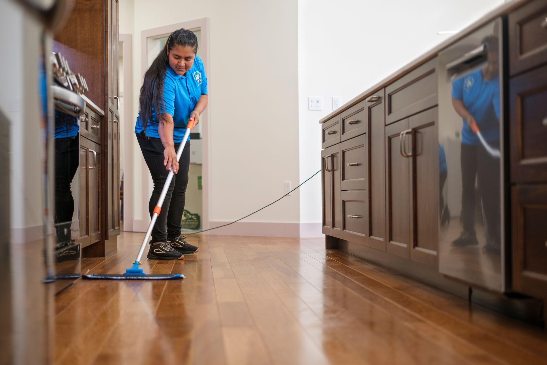 Person mopping hardwood floor in a kitchen, wearing blue shirt and black pants.