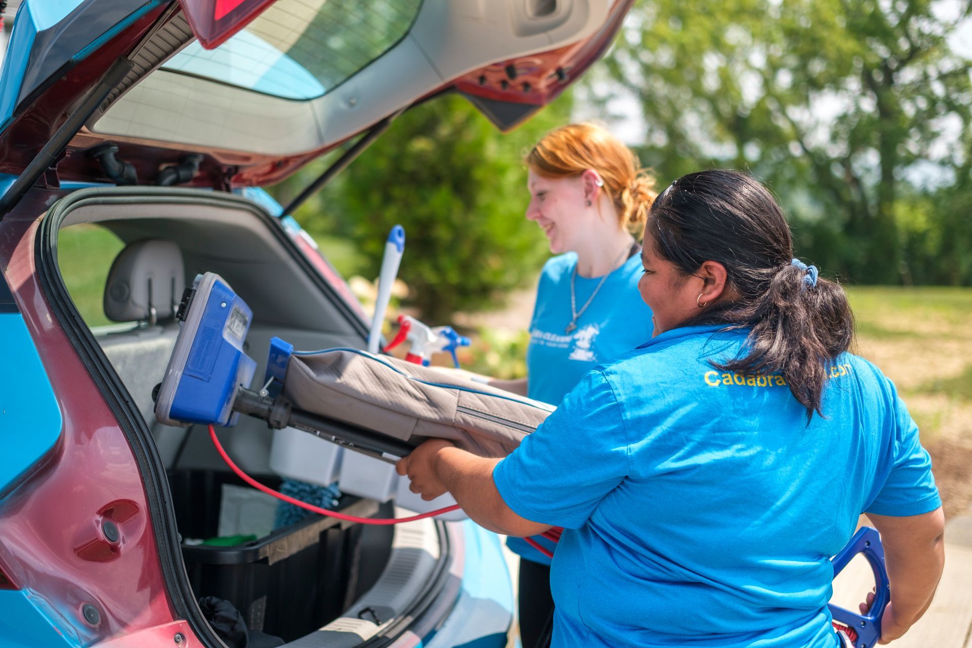 Two people load supplies into a car's open trunk. One person wearing blue removes a cleaning device. Sunny outdoors.