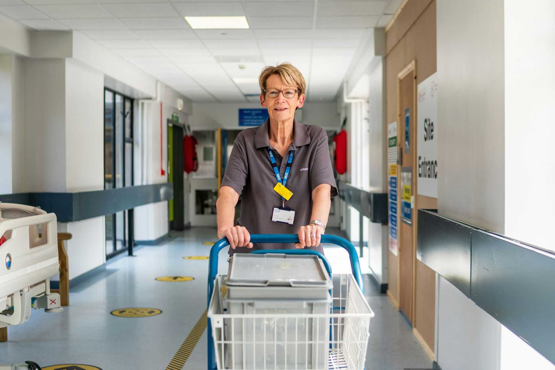 A person in a hospital corridor pushing a cart with a grey storage bin.