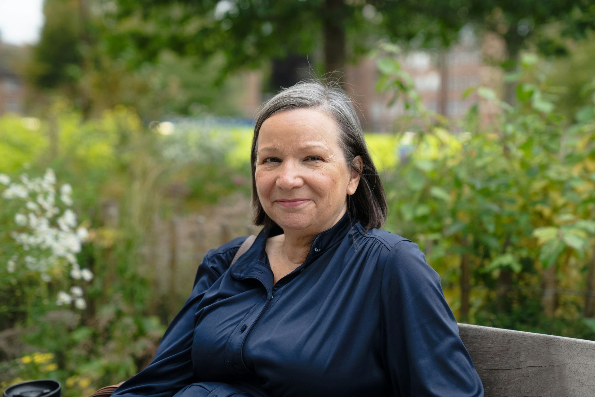 A person with grey hair wearing a navy shirt sits on a bench outdoors in a garden setting, smiling at the camera.