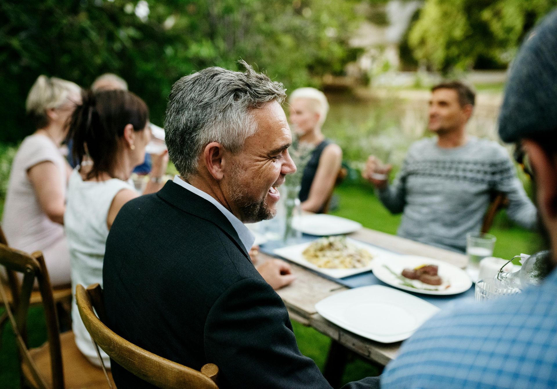 A group of people having an outdoor dinner, focusing on a smiling man in a suit sitting at a table with food.