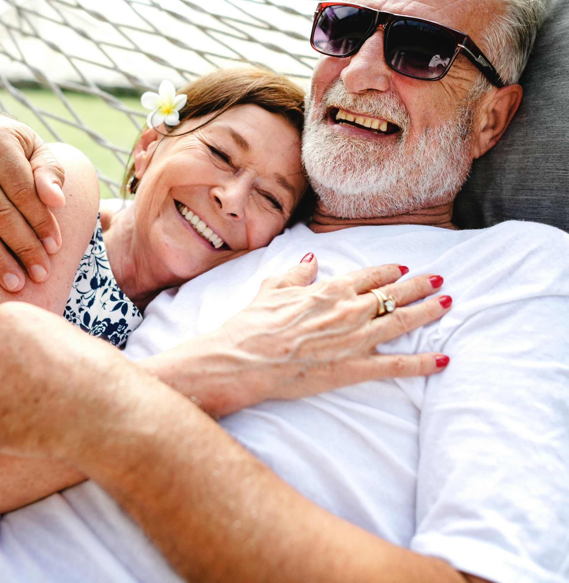 A person with a flower in their hair embracing another person wearing sunglasses while relaxing in a hammock.
