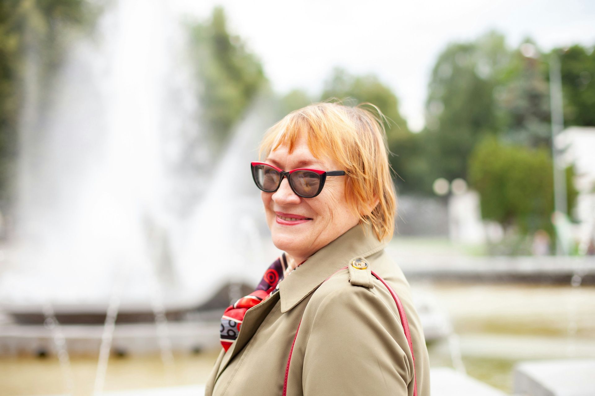 A person with blonde hair and glasses wearing a khaki trench coat stands smiling in front of a blurry fountain.