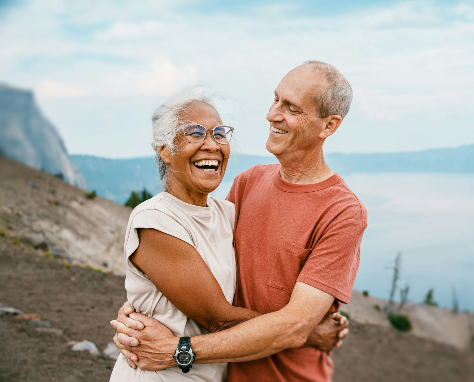 Two people embracing outdoors near a lake and mountains, smiling at each other.