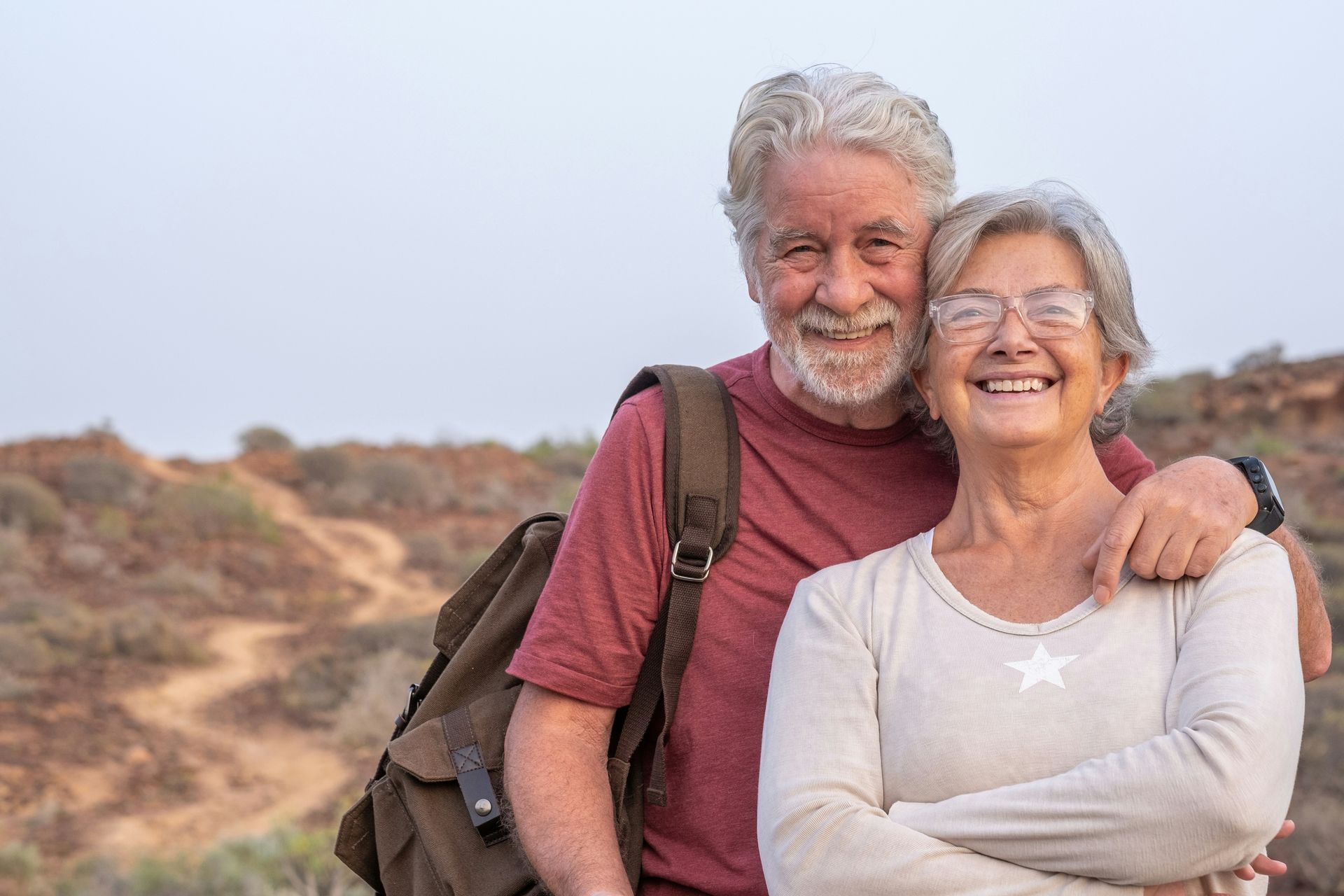 A smiling couple with backpacks standing outdoors in a dry, desert landscape.