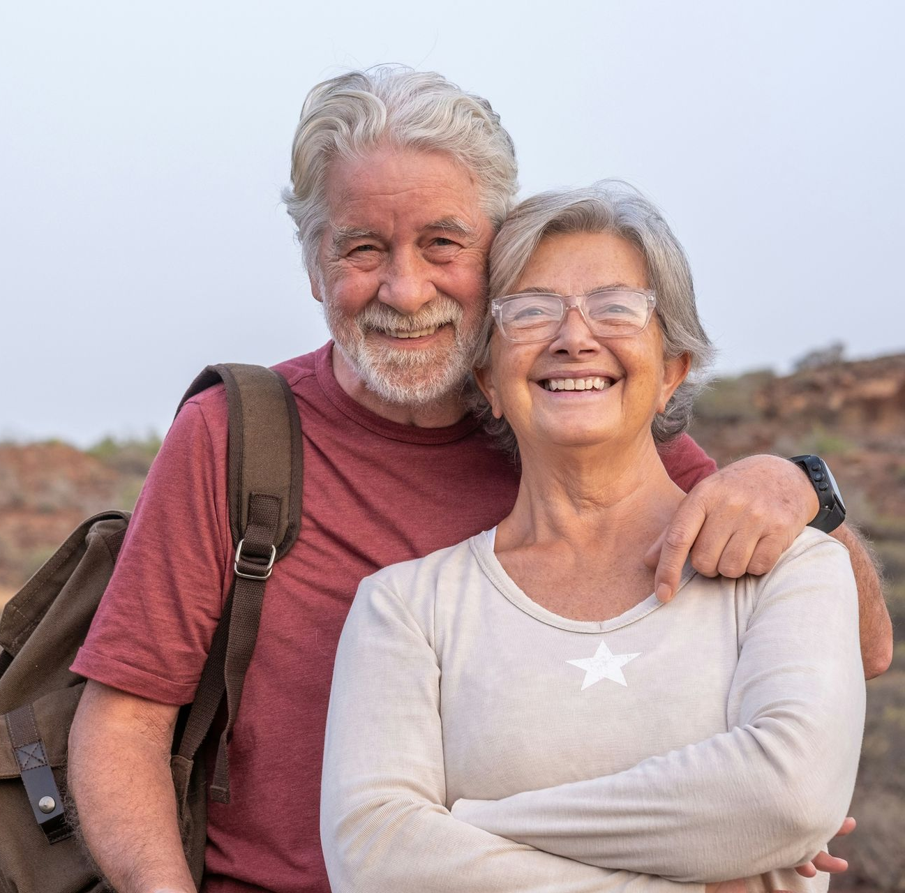 Two people embrace outdoors, smiling at the camera. The person on the left wears a maroon t-shirt and a brown backpack.