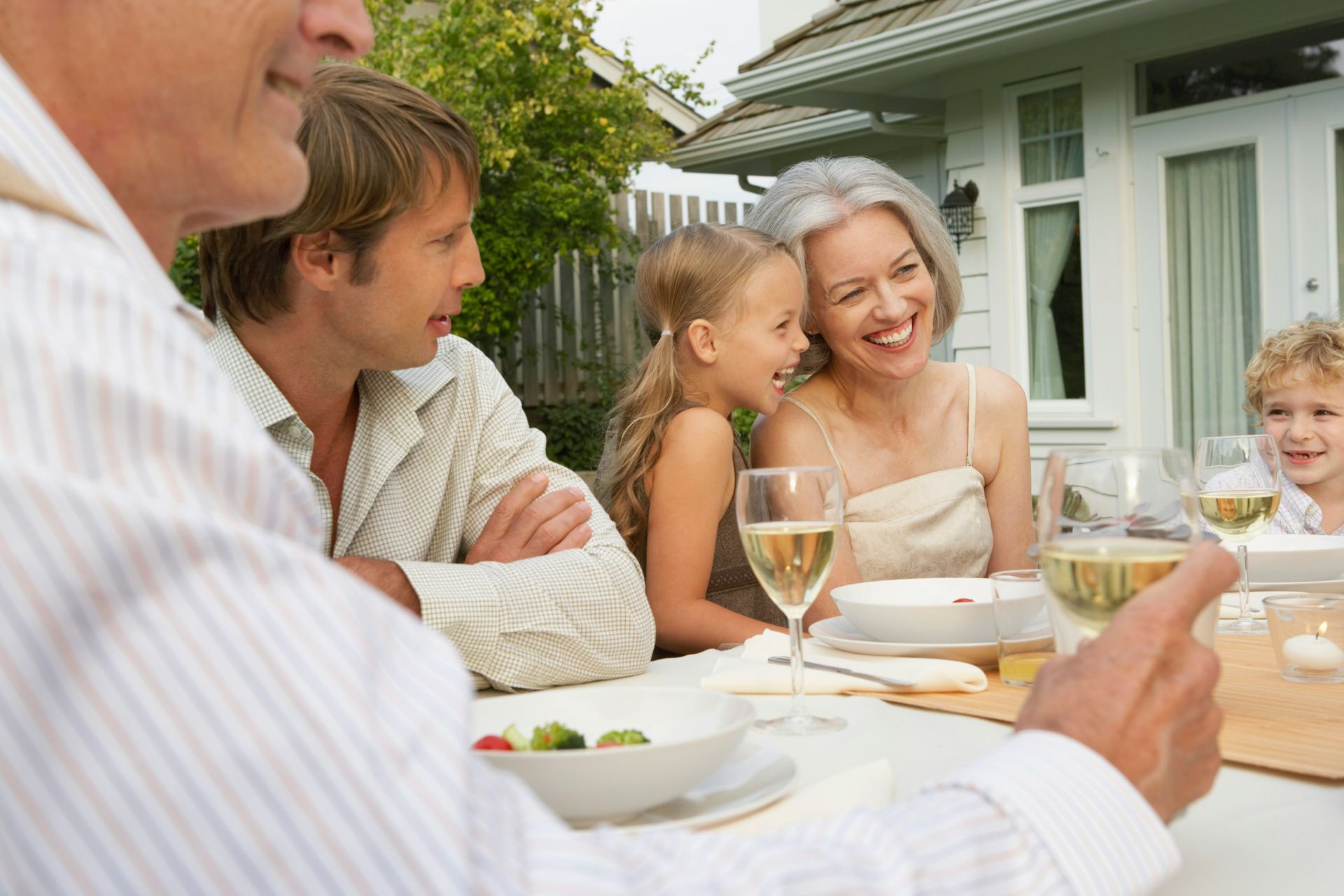 A multigenerational family enjoying an outdoor meal, laughing and holding glasses of white wine at a patio table.