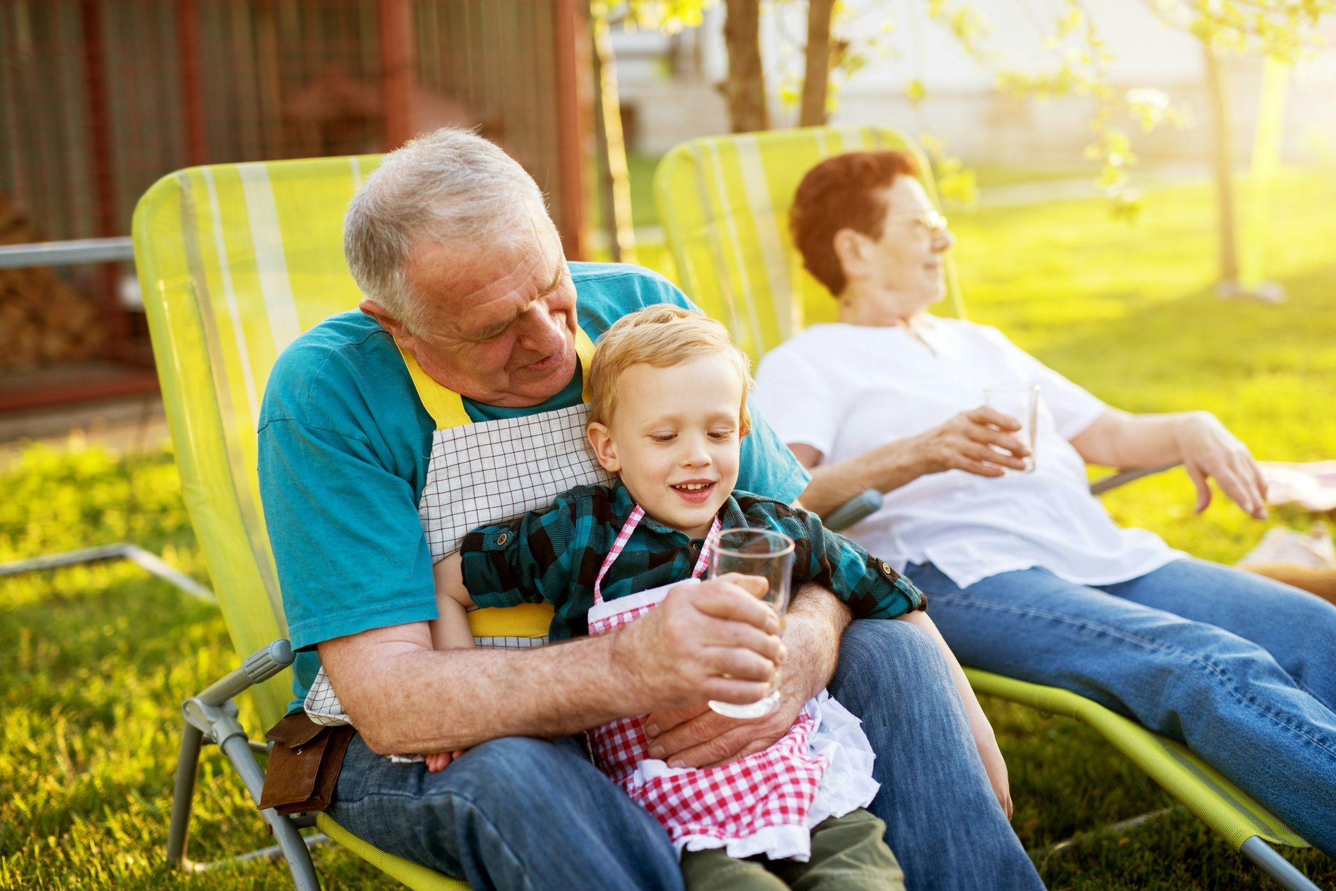 A child sits on a person's lap, holding a glass, with a second person relaxing in a lawn chair in a sunny garden.