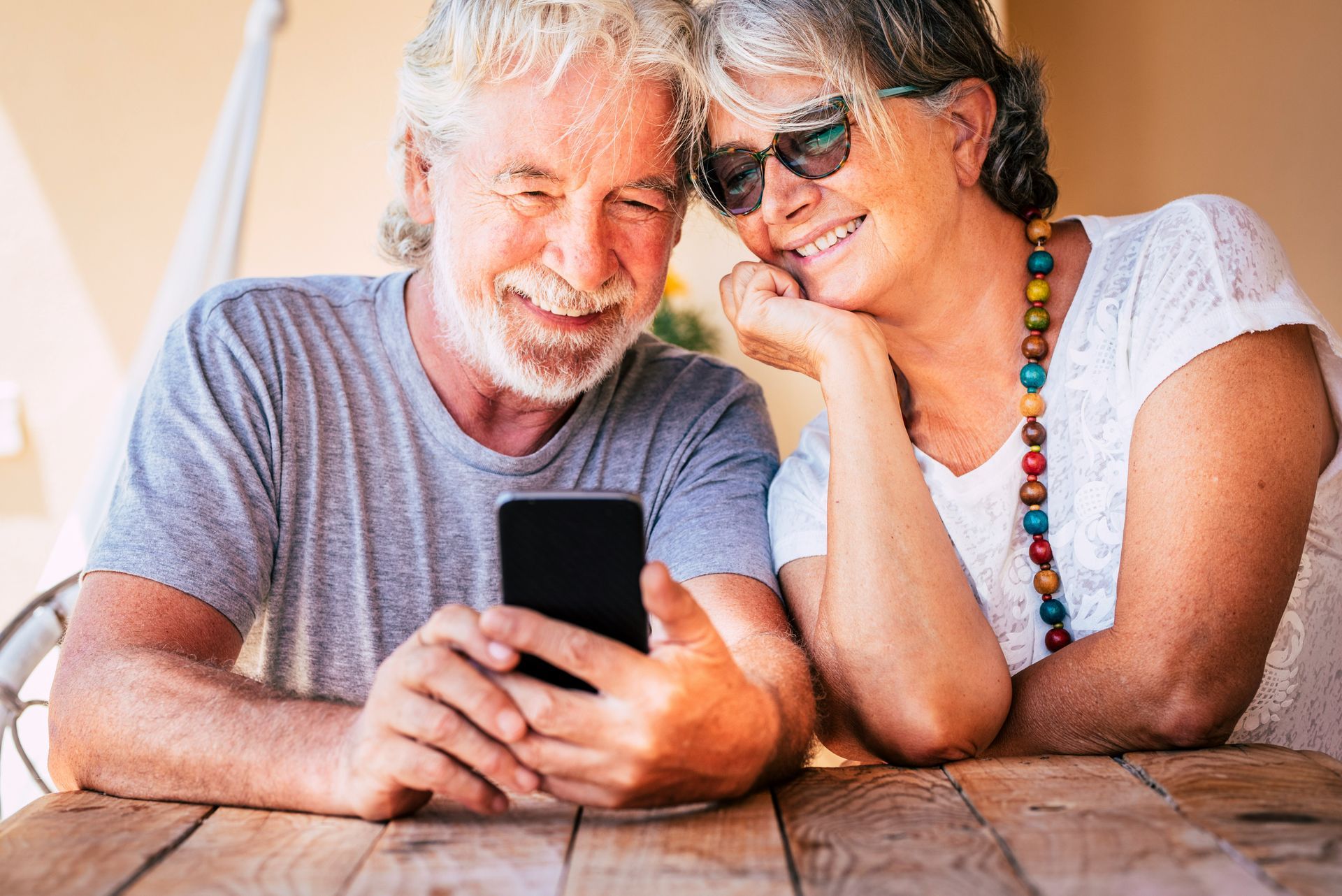 Two people sit at a wooden table, smiling and looking down at a smartphone together.