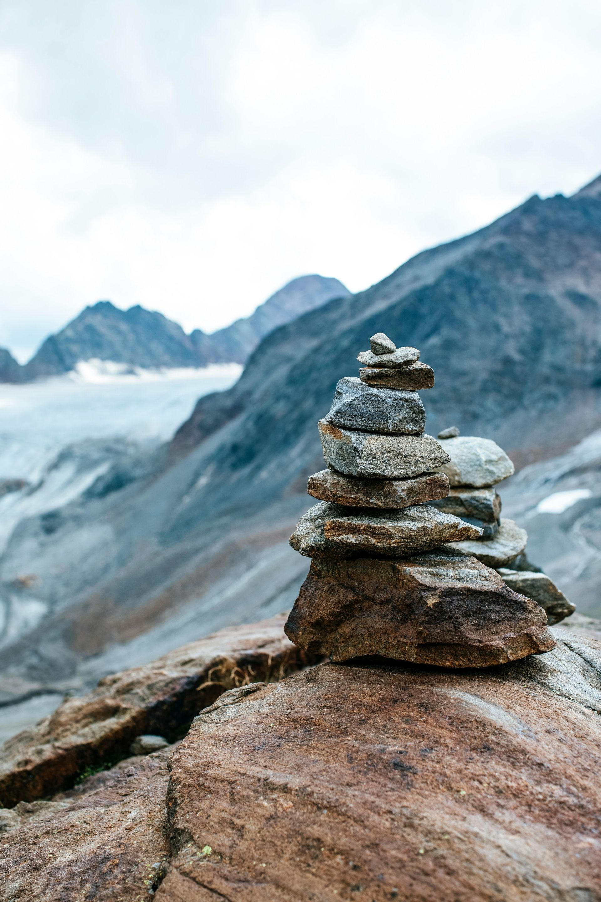 A small stack of balanced rocks sits on a cliff overlooking a vast, snow-covered mountain range.