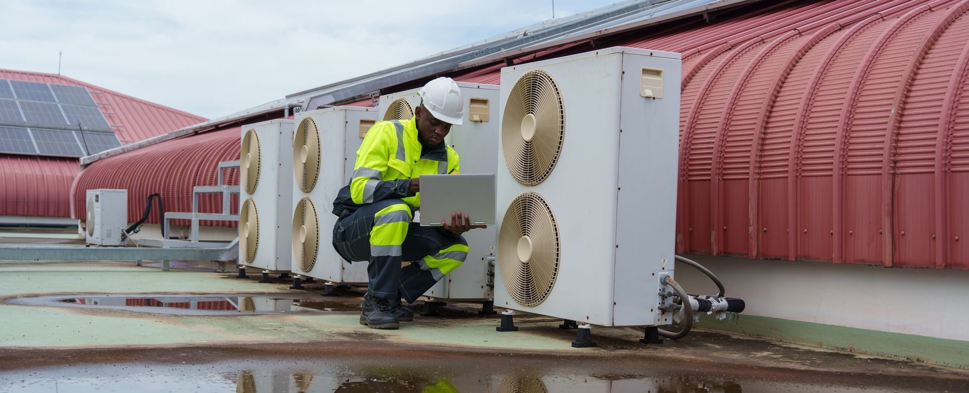 Worker in yellow safety gear inspecting rooftop HVAC units beside a red curved building. Worker in yellow safety gear inspecting rooftop HVAC units beside a red curved building.