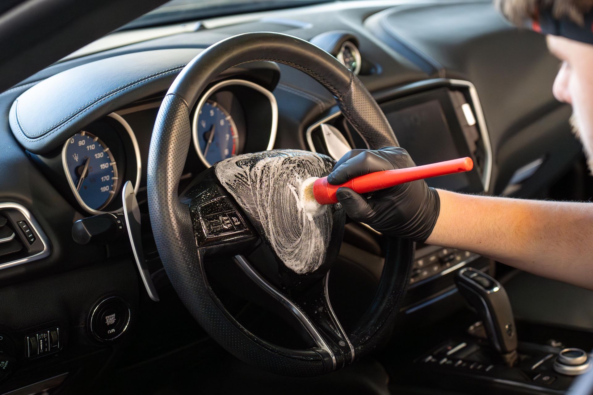 Person wearing gloves cleaning a car's steering wheel with a brush and cleaning product.