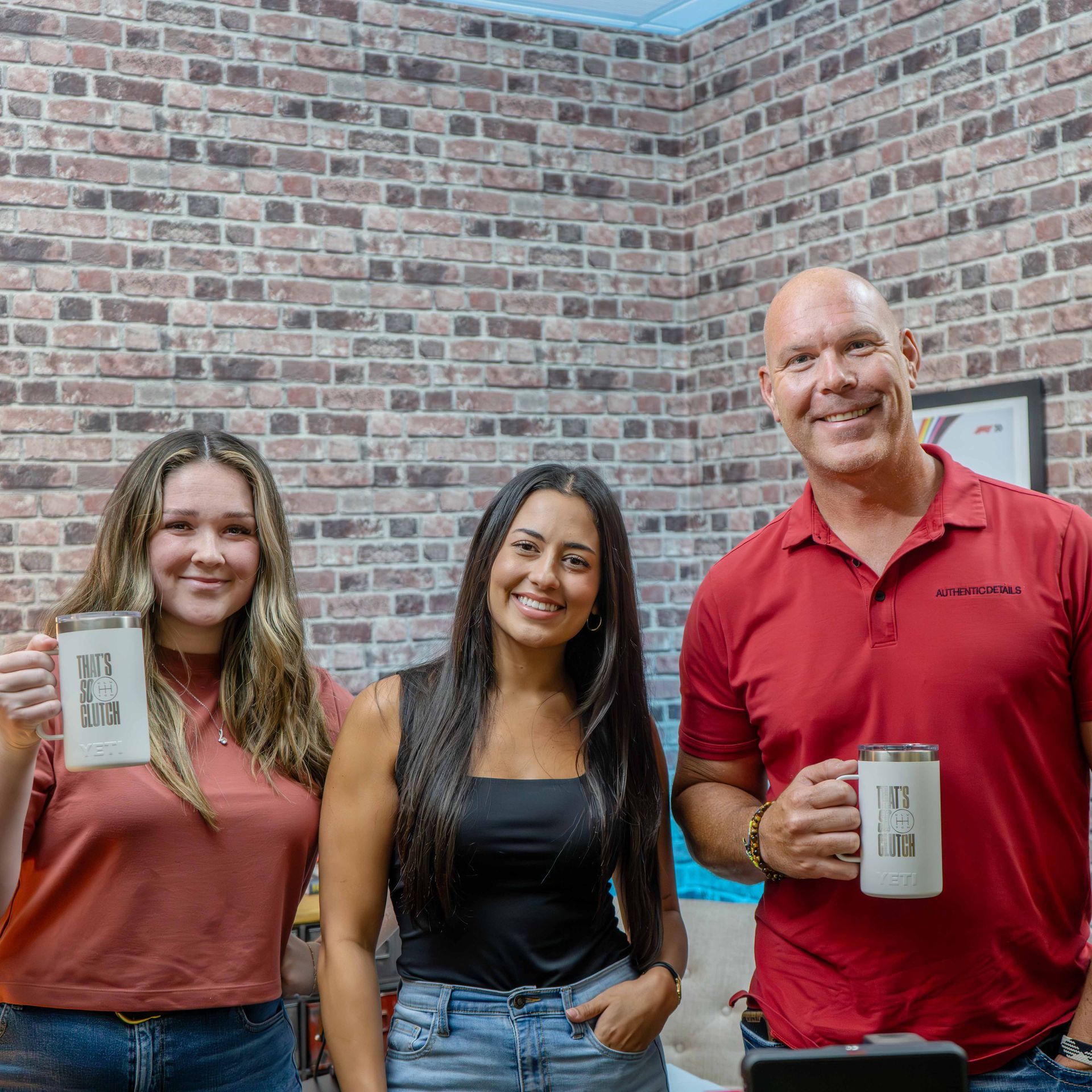 A man and two women are standing next to each other holding coffee mugs in front of a brick wall.