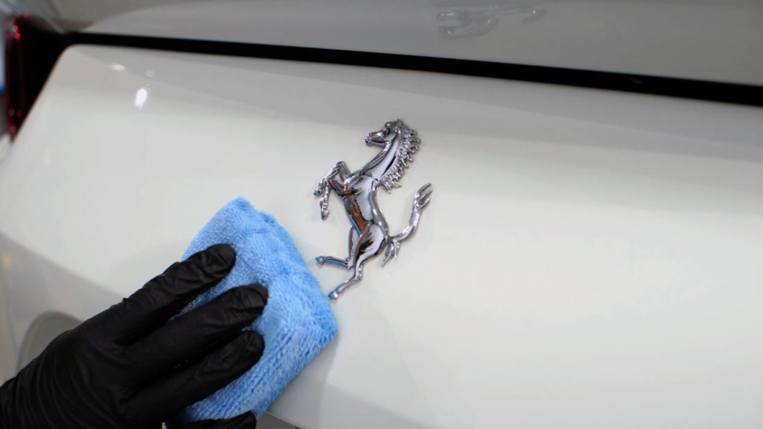 Person in black gloves cleaning a white car with a blue cloth, focusing on the chrome Ferrari emblem.