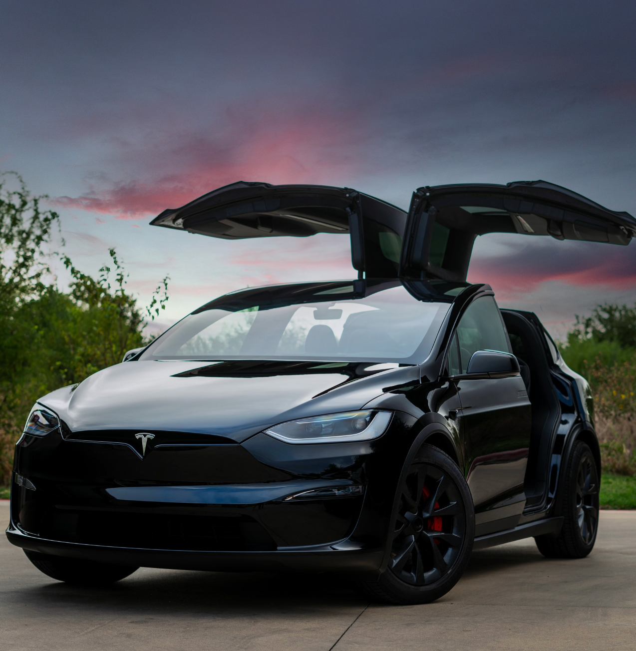 Black Tesla Model X SUV with falcon wing doors open against a sunset sky.